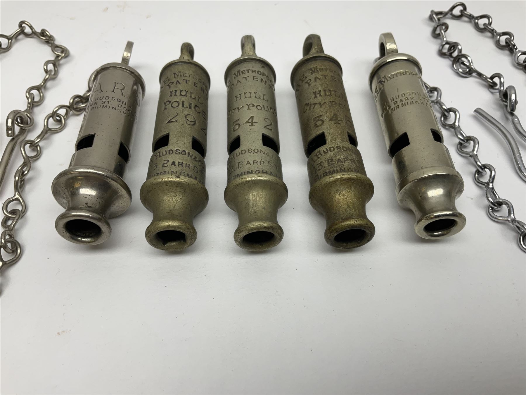 Three Hull City Police 'Metropolitan' whistles by J. Hudson 13 Barr Street Birminham, each with impressed collar number; another 'Metropolitan' police whistle with chain; and an ARP whistle by Hudson & Co with chain (5)