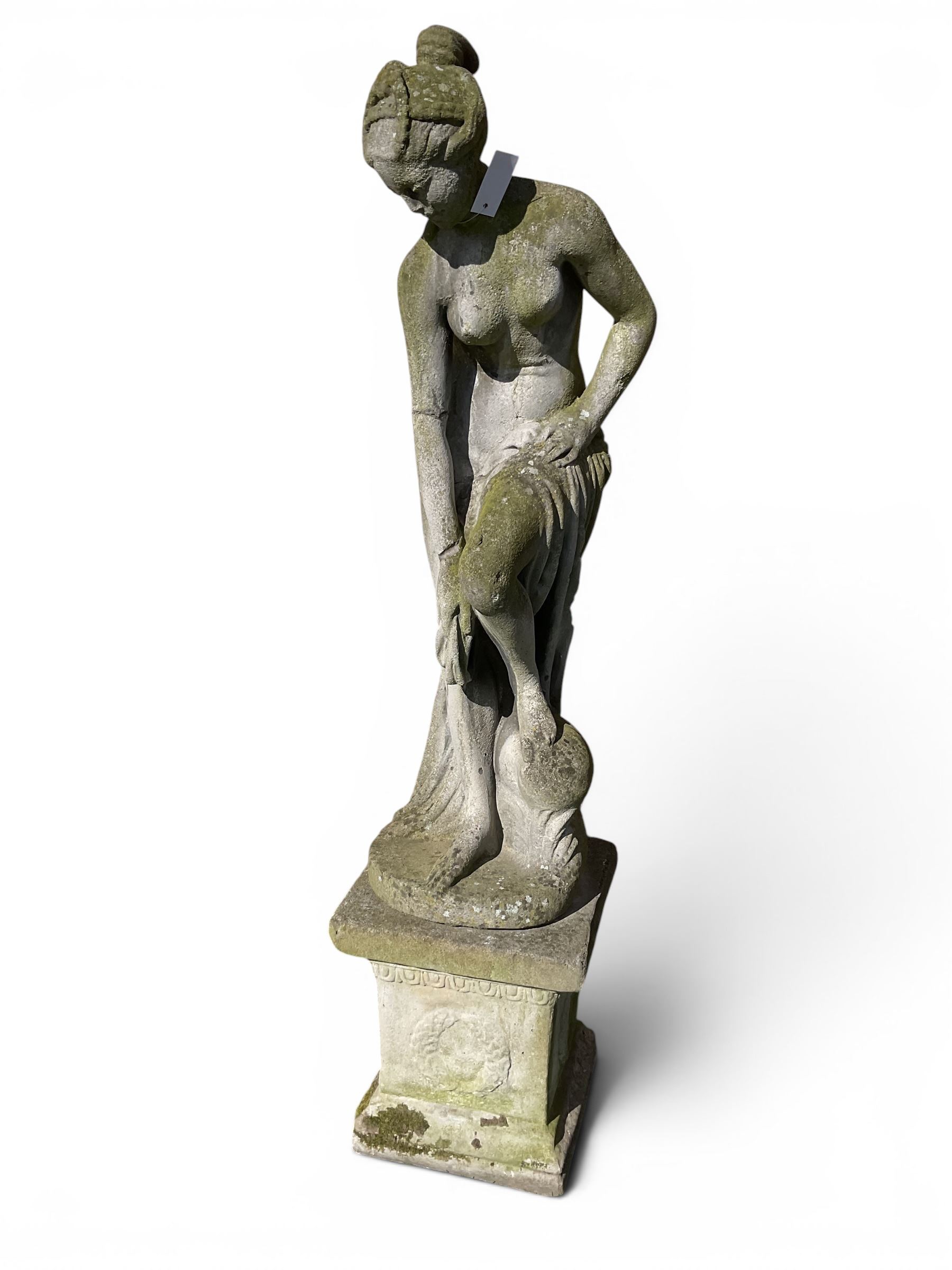 Cast stone garden figure of a classically draped nude female inspired by Aphrodite statues, with stylised up-do arranging her chitin on a circular base, the rectangular pedestal with egg-and-dart moulding over a floral wreath, in weathered finish