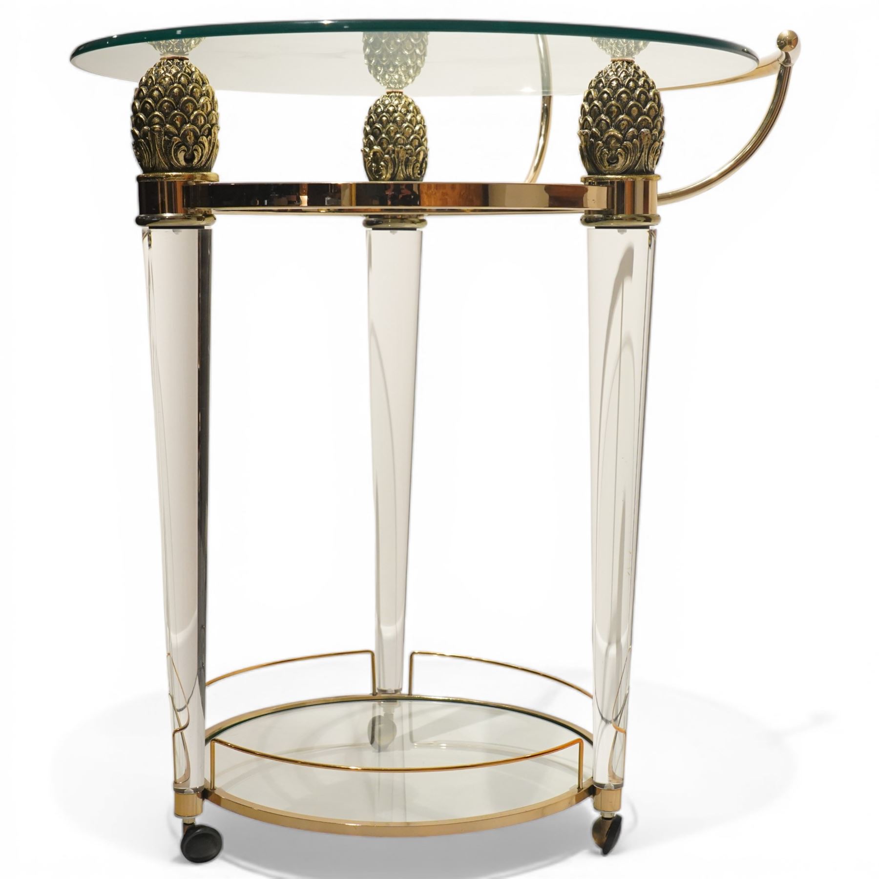 Hollywood Regency style gilt metal and glass serving trolley, circular top in a polished brass surround with three pineapple finials, supported on clear column legs, lower glass shelf enclosed by a brass gallery, terminating in castors