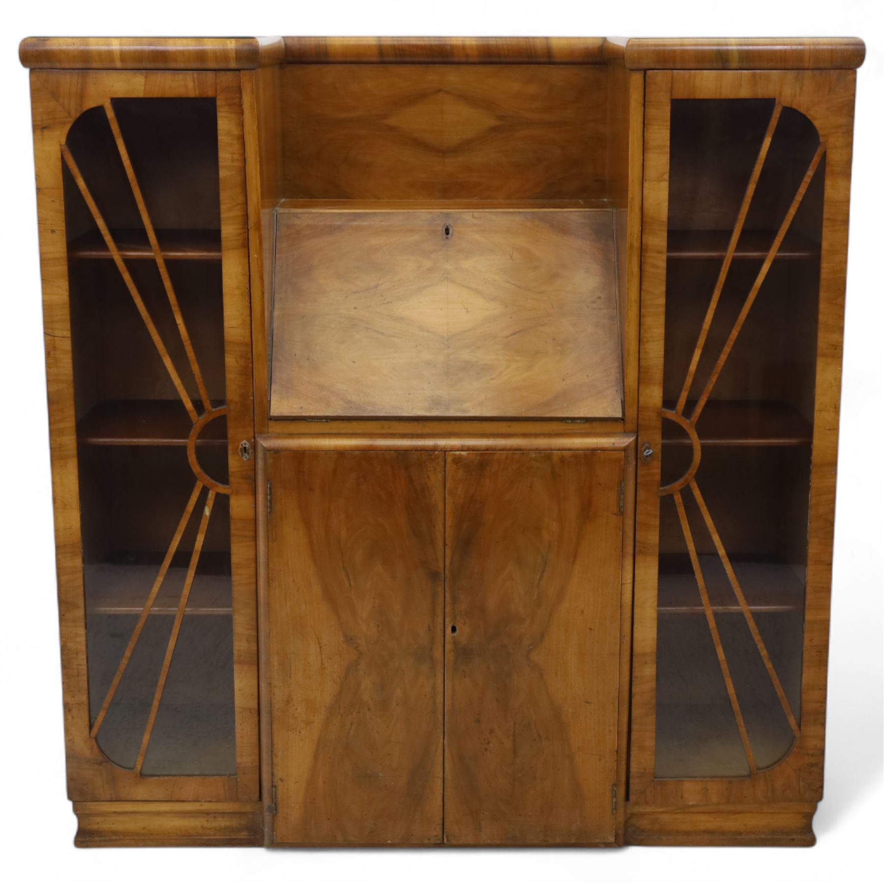 Art Deco walnut bureau bookcase, top with moulded and rounded edges and stepped front profile, central shelf, and fall-front bureau section with vertical and horizontal pigeon holes and a small drawer, above two cupboard doors and flanked by two tall glazed display cabinets with single doors and geometric sunburst design astragal glazing bars enclosing three shelves, raised on cavetto shaped plinth base 
