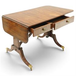 Early 20th century Regency Revival figured mahogany sofa table, rectangular drop-leaf top with rounded corners, fitted with two drawers to each side with brass flowerhead pull handles, raised on twin end supports with splayed reeded legs and brass cups and castors, united by a scrolling and reeded closed stretcher