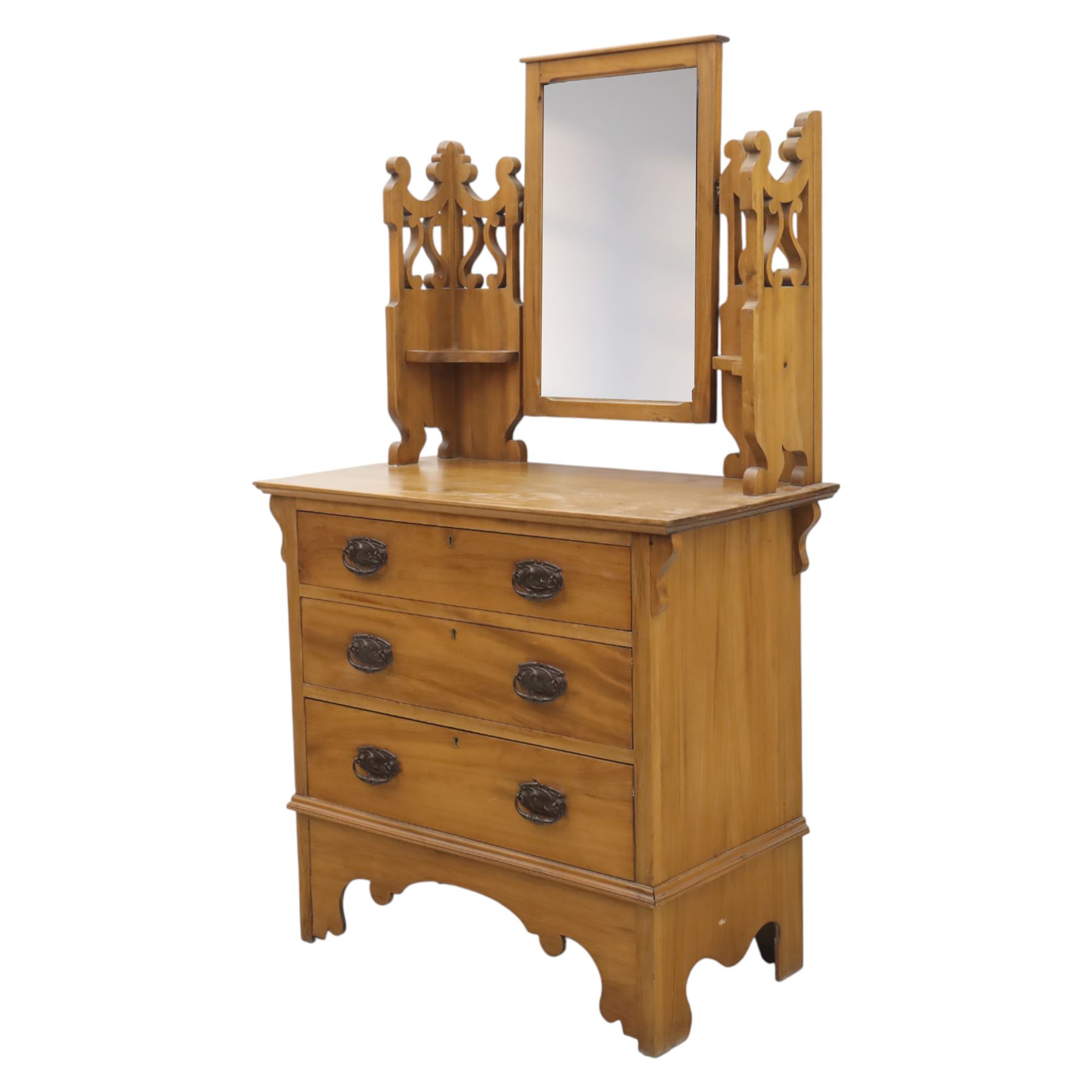 Edwardian stained pine dressing chest, the moulded top over three graduating drawers with pressed brass handles, shaped apron and bracket feet, the mirror back with rectangular plate on pivot supports, flanked by pierced Gothic style uprights with small corner shelves