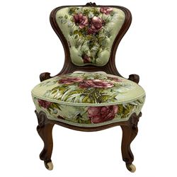 Victorian walnut framed nursing chair, spade shaped back carved with flower heads, upholstered in buttoned rose patterned fabric, on scroll carved cabriole supports terminating at brass and ceramic castors