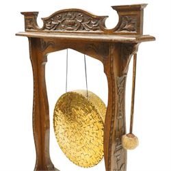 Late Victorian Arts & Crafts carved oak framed floor standing dinner gong, the cresting rail carved with central urn and trailing foliage, over shaped end supports decorated with graduating tulip and floral motifs, on rectangular base with scroll sledge feet, the brass gong stamped with William Suckling of Birmingham 'Burmese' makers mark, with striker