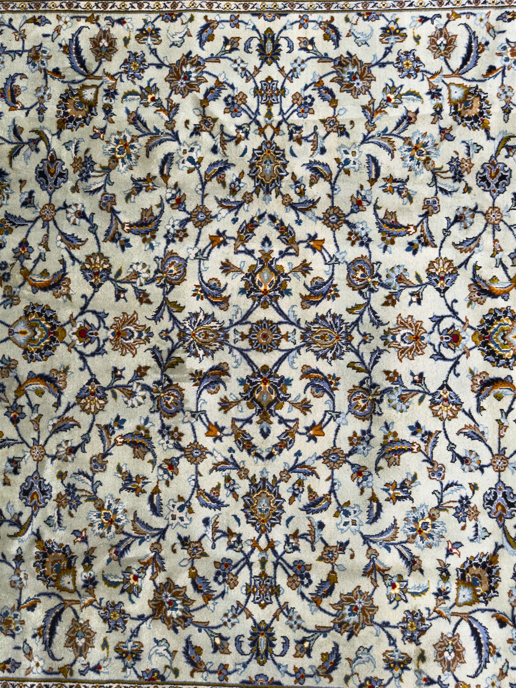 Central Persian Kashan hand-knotted carpet, the ivory ground intricately decorated with scrolling vines, palmettes, and stylised flowerheads in soft blue, gold and brown tones, enclosed by a broad indigo border with repeating floral motifs and multiple guard stripes