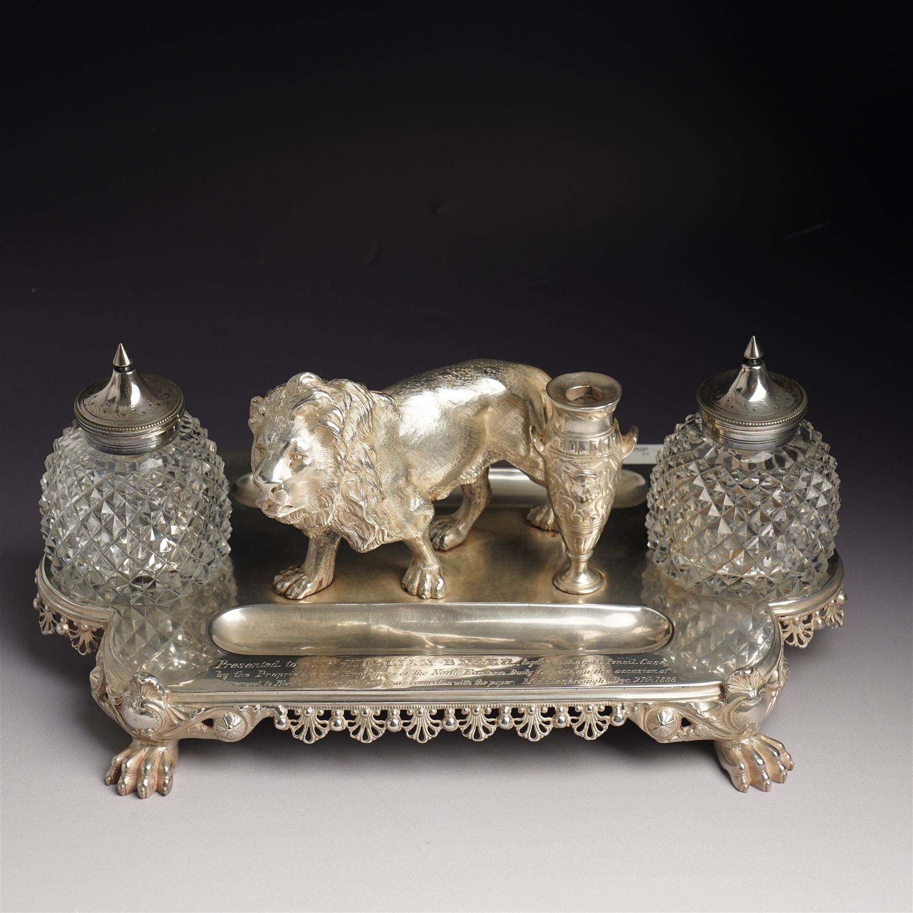 19th century Walker & Hall, silver plated inkstand, oblong and on four paw feet, the border decorated with pierced shell design, each side with a pen-rest, with two cut-glass bottles and central lion and urn finials, engraved to the front with an inscription, H15cm, L30cm