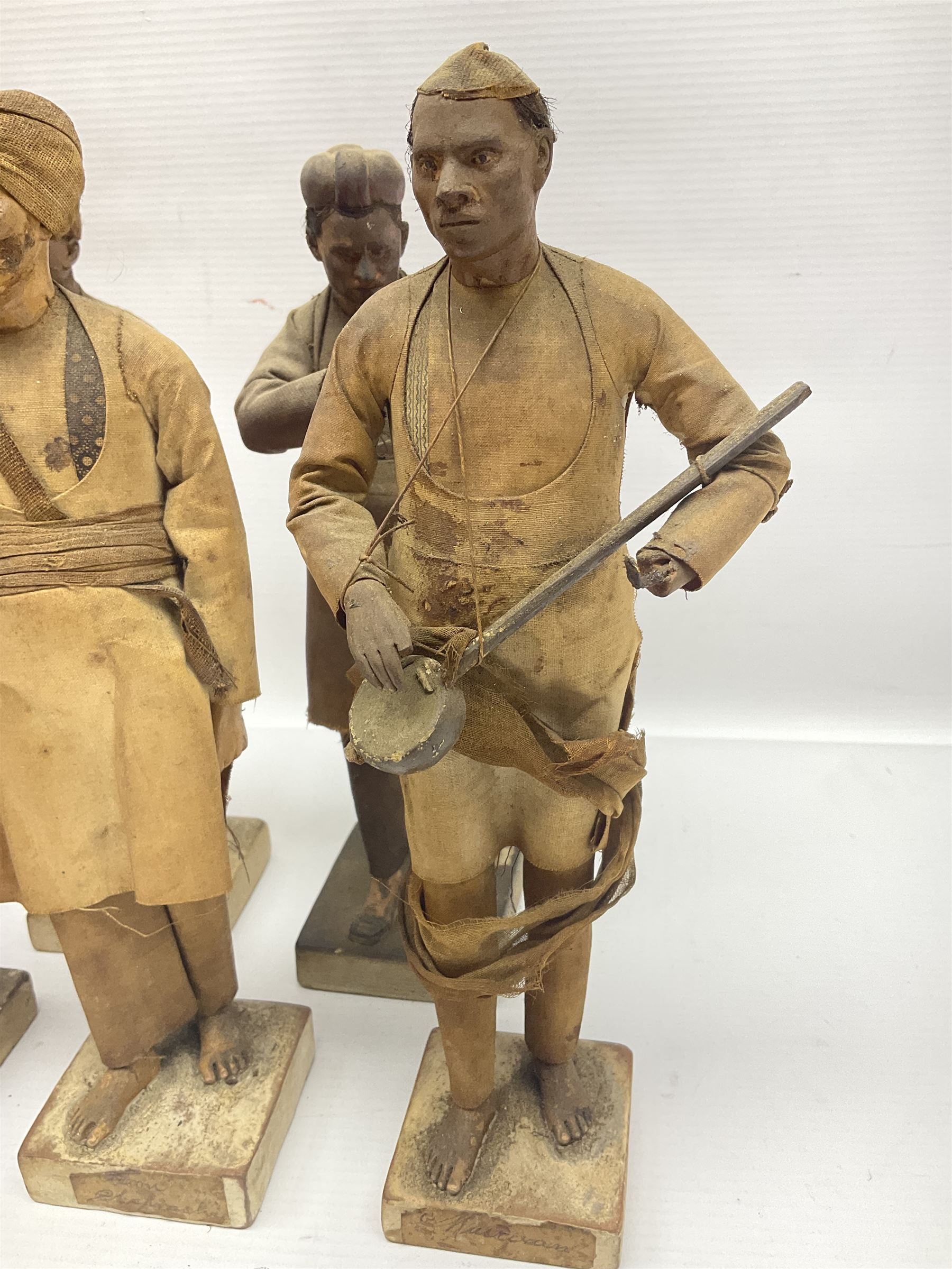 Seven 19th Century Indian clay dolls, possibly Krishnanagar, each figure with painted details, wearing cloth garments, standing on rectangular bases, some with paper label to the base, H25cm