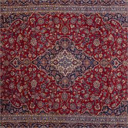 Central Persian Kashan hand-knotted carpet, madder red field with central cusped medallion in navy and ivory enclosing a floral rosette and radiating scrolling arabesques and palmettes, across a densely ornamented ground of finely drawn vinery enriched with a profusion of flowering sprays and stylized blossoms, spandrels in deep indigo and principal border of navy ground decorated with a continuous palmette and vine-scroll flanked by multiple guard stripes, with cotton fringe ends