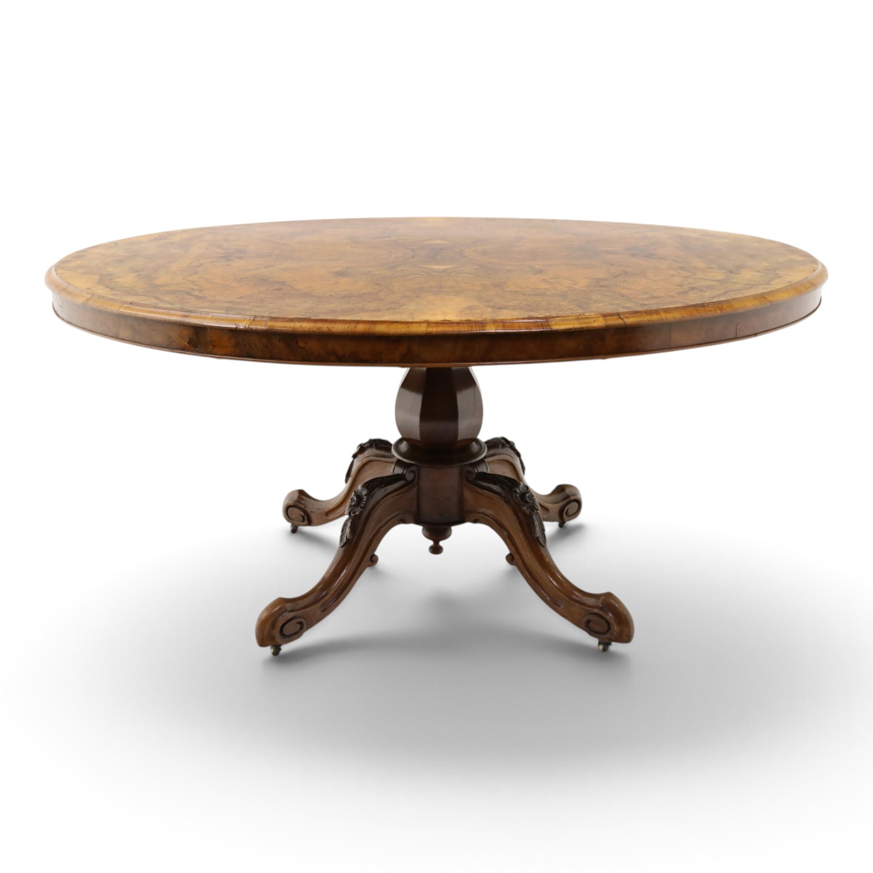 Victorian figured and burr walnut loo table, oval book-matched veneer top with moulded edge, over a turned octagonal pedestal terminating to four splayed supports with flowerhead carved knees and scroll feet with castors
