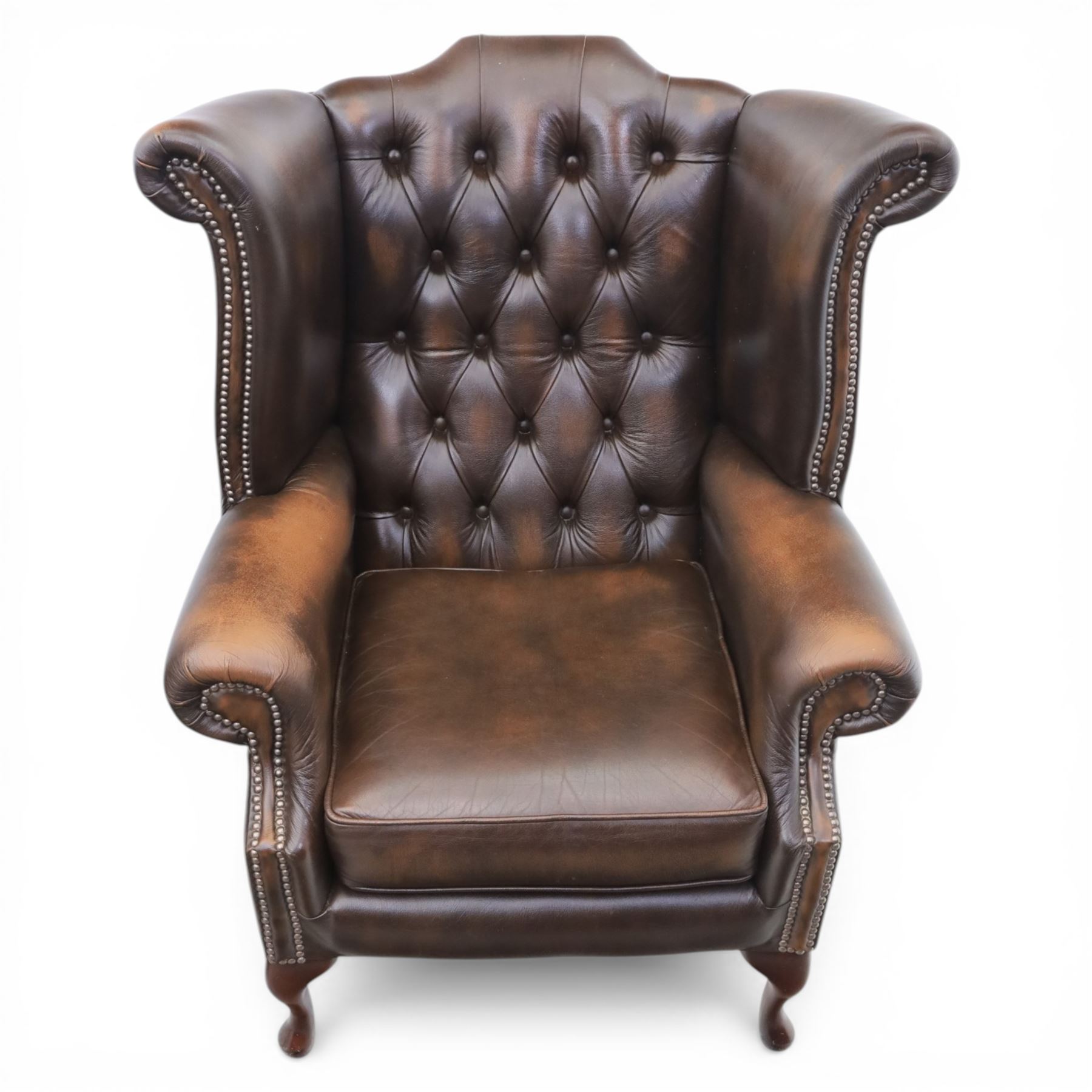 20th century Chesterfield style brown leather wingback armchairs, with button upholstered backs and wings, shaped scroll arms with studded borders, loose seat cushions, raised on cabriole supports with pad feet