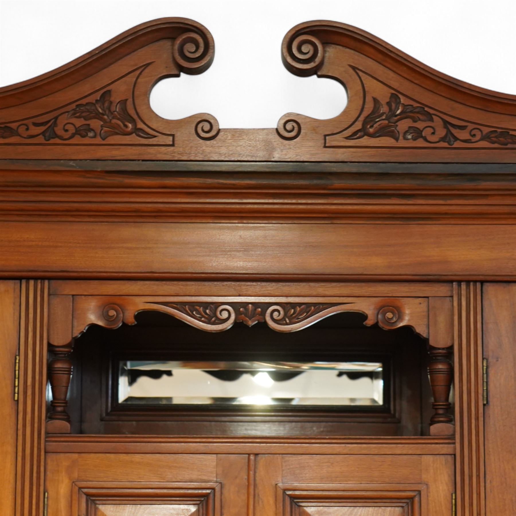 Edwardian walnut triple combination wardrobe, projecting pediment with pierced foliate scroll cresting above a central cupboard flanked by bevelled mirrored doors, the central section with twin panelled cupboard doors carved with foliate scrolls, over open recess with mirrored back and turned supports, above two short and three long drawers with brass handles, interior fitted with hanging rails and shelf, raised on plinth base
