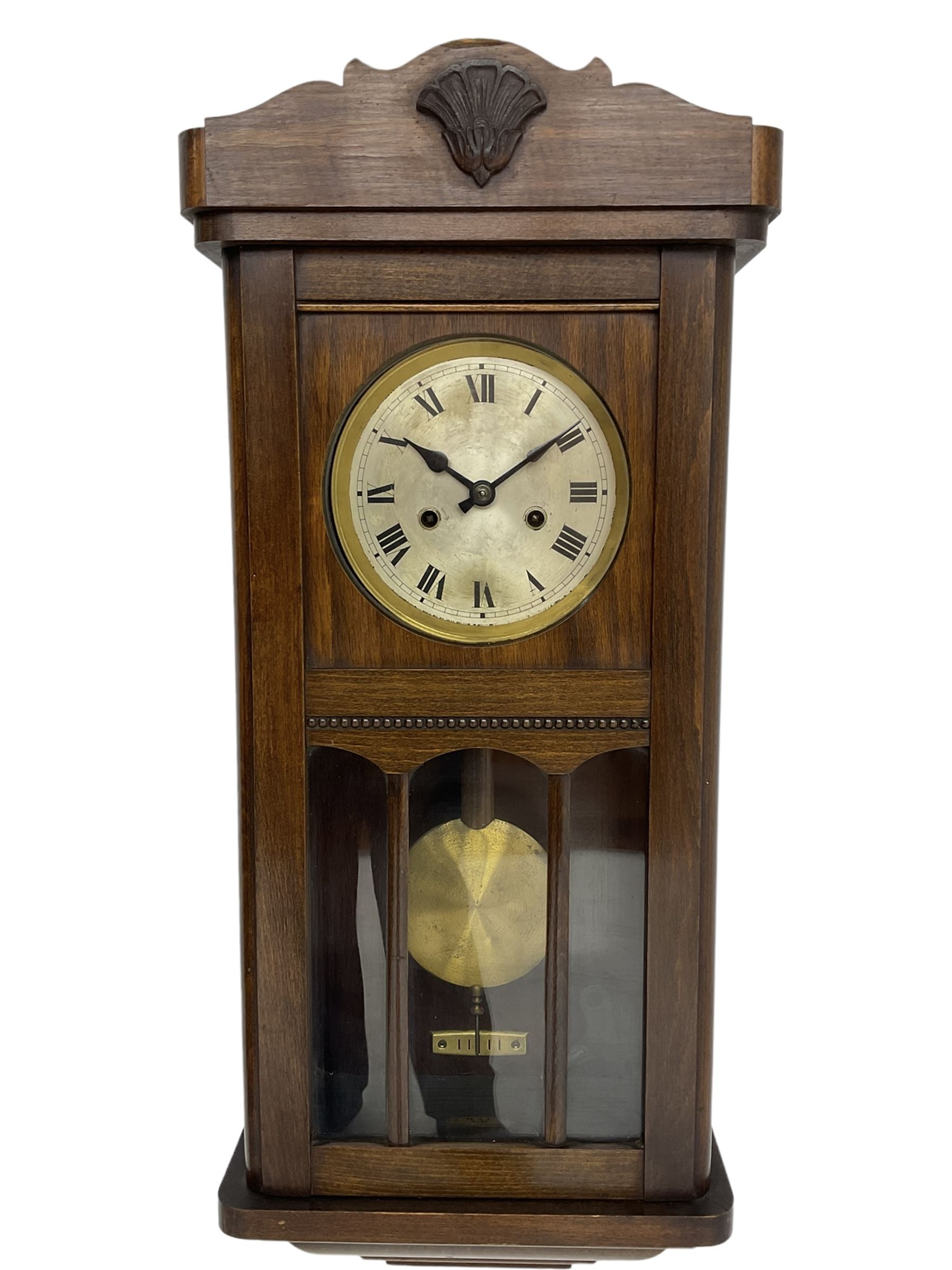 1930s wall clock in an oak case with a crested pediment and glazed door with visible pendulum, silvered dial with Roman numerals and steel spade hands, eight day spring driven movement striking the hours and half hours on a gong. With pendulum and key.