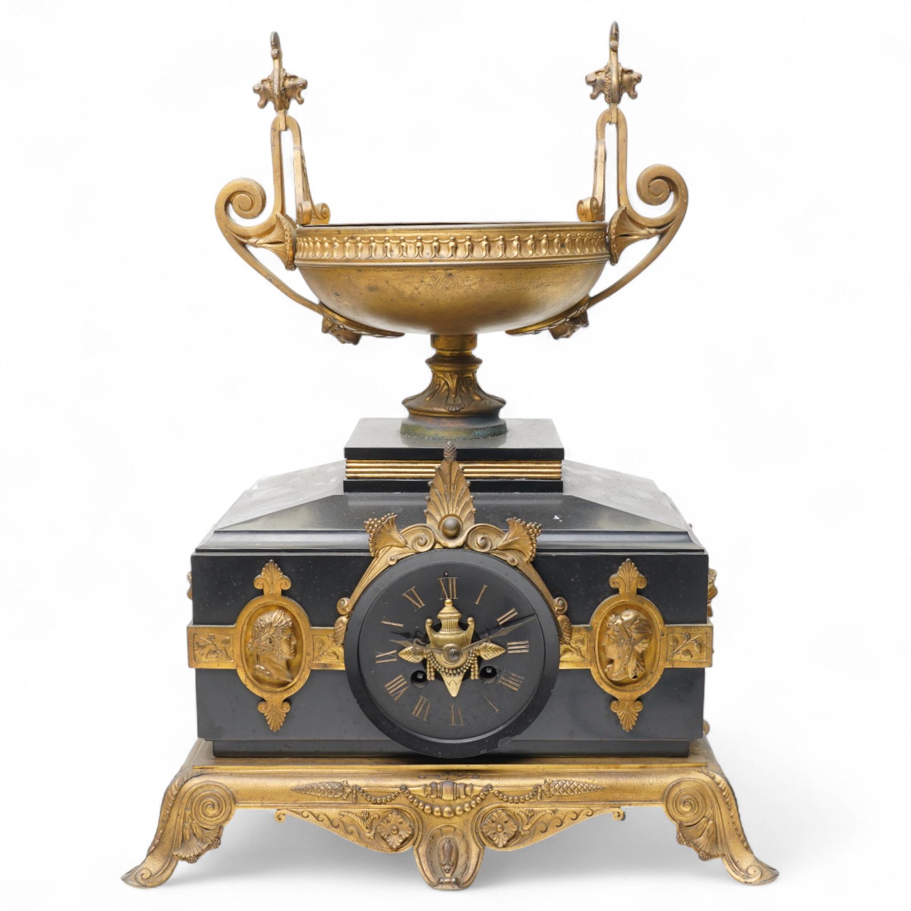 French- late 19th century 8-day mantle clock in an imposing Belgium slate case with gilt metal ornamentation. Stepped rectangular case with an angled top and square plinth surmounted by an oval shaped gilt urn with scroll handles, case with continuous strapwork and oval medallions displaying portraits from classical antiquity, raised on a gilt metal base with splayed feet, conforming unenclosed slate dial with gold incised Roman numerals and brass Fleur de Lis hands, Parisian twin train rack striking movement with pendulum regulation, striking the hours and half hours on a bell. With pendulum.