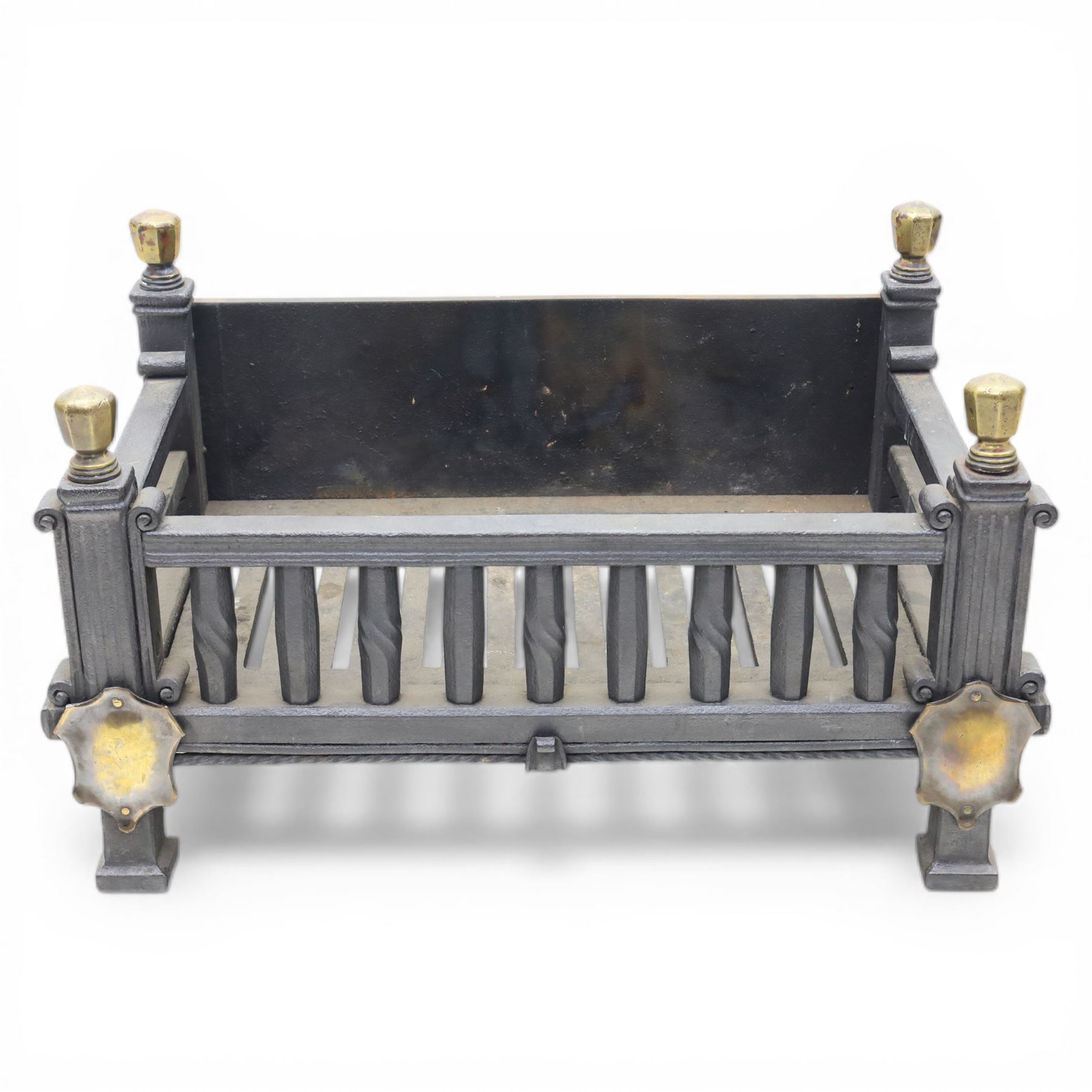 Arts and Crafts cast iron fire grate, rectangular backplate with plain upper rail over a slatted front of cylindrical tapering bars, flanked by square section uprights with fluted detail and scroll terminals, raised on stepped feet with shaped brass roundel mounts and brass finials to the corner posts