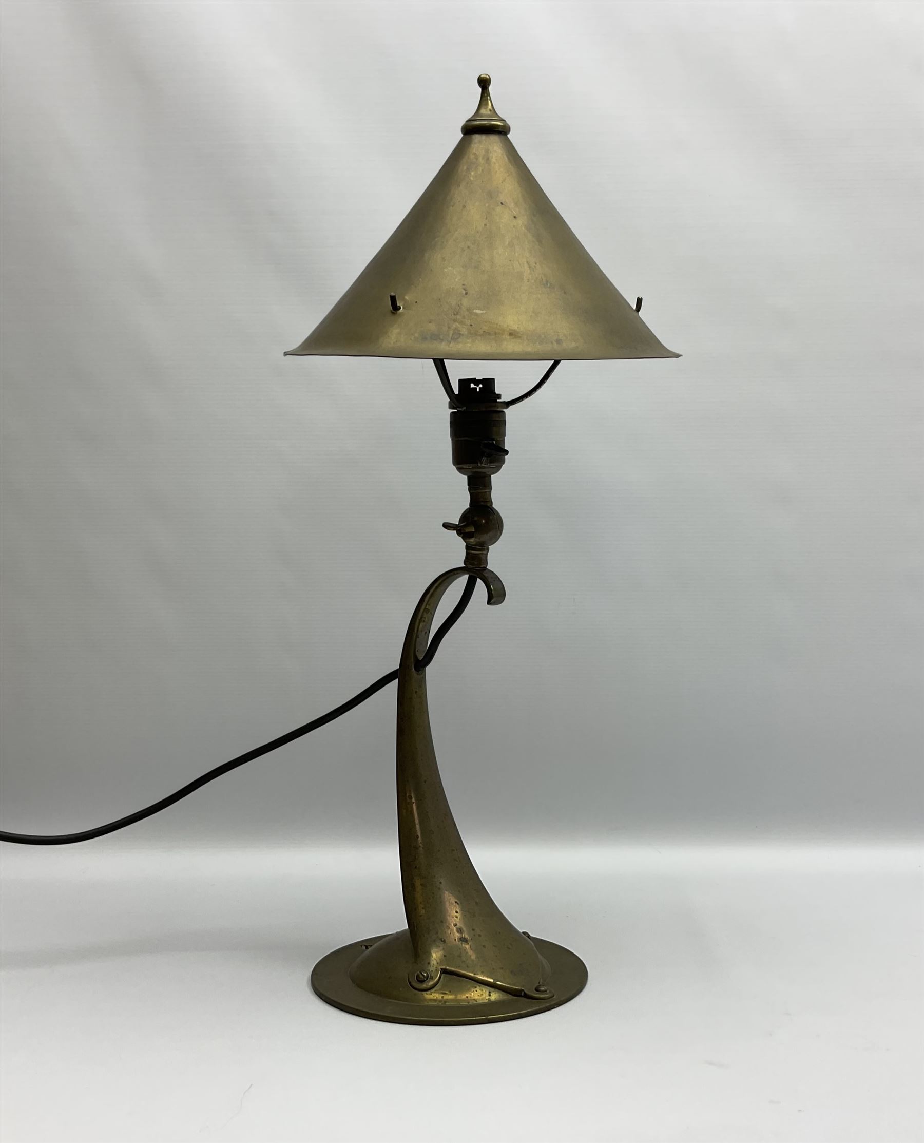 Arts & Crafts Jesson, Birkett & Co, brass table lamp or wall light, designed by Thomas Birkett, the tapering stem with adjustable ball pivot supporting a conical shade with tapering final with ball terminal, upon a circular domed base pierced for wall mounting, shade positioned upright overall H57cm

Often misattributed to Arthur Stansfield Dixon for Birmingham Guild of Handicraft, this particular lamp was designed by Thomas Birkett.
In the late 1890's Birkett worked as a metalwork at the Birmingham Guild, before later joining the Faulkner Bronze Company which went on to become Jesson, Birkett & Co in 1904. The lamp features in lighting catalogues from the firm under the model no E490.


