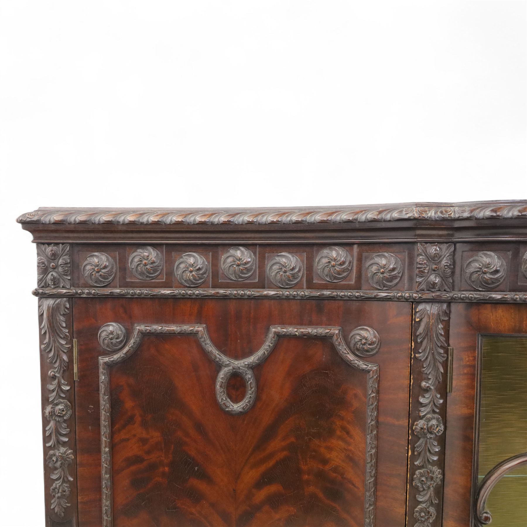 Edwardian mahogany serpentine breakfront display cabinet in the neoclassical style, the rectangular top above a carved frieze with roundels, foliage, and ribbon tied swags with central bow-fronted astragal glazed doors enclosing shelves, flanked by panelled doors, raised on bracket feet 