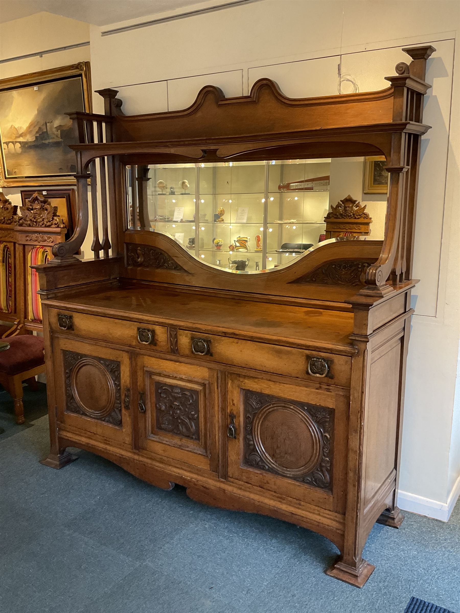 Late 19th century Arts & Crafts period oak mirror back sideboard, shaped bevelled mirror back carved with foliate scrolls and flower heads, down swept supports with elongated club-shaped uprights, moulded rectangular top over two drawers and three cupboards, the drawer fronts with copper handles decorated with sailing ships, panelled doors carved with mythical beasts, flowerheads and foliage, on moulded block feet