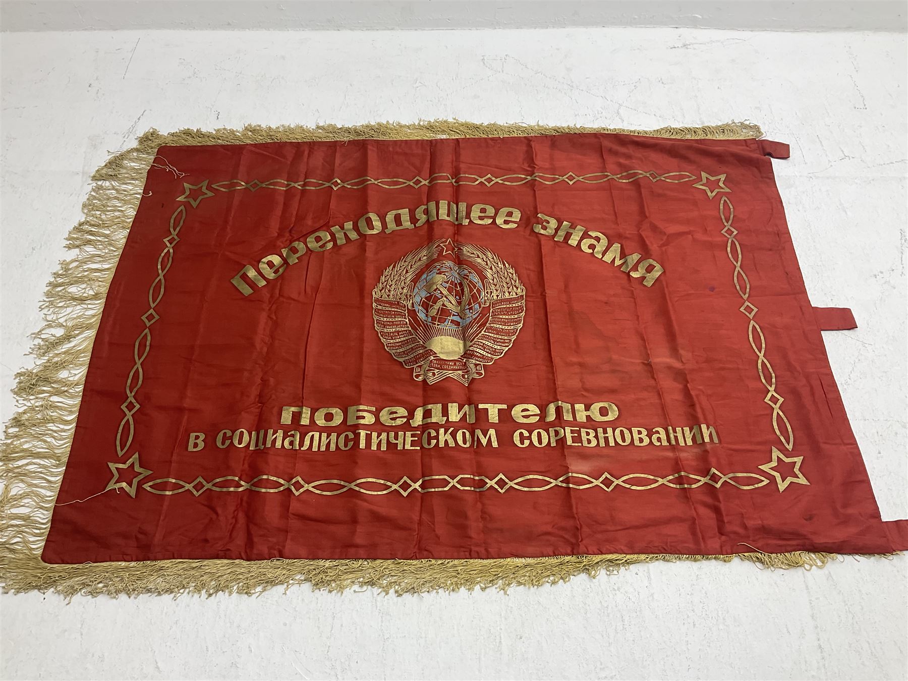 1970s Soviet banner printed in gold on a red ground, roughly translates as 'Transferable Banner - To the winner in the Socialist Competition' and 'Work Study Live by Communism' verso; wreath of wheat ears to either side; tassels on three sides 110 x 155cm