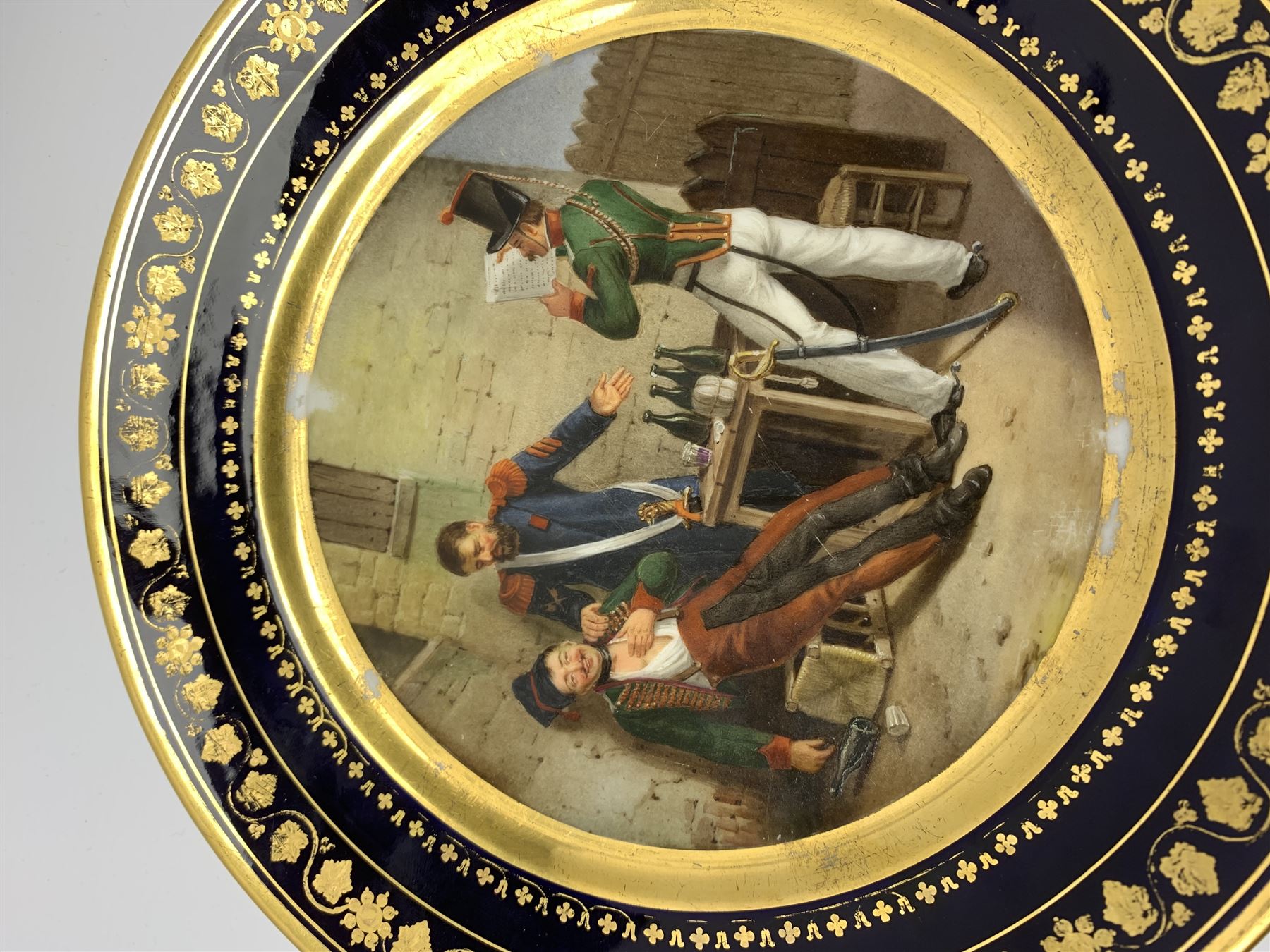 19th century Rihouet Paris porcelain cabinet plate, decorated with a central panel illustrating three French soldiers around a bottle laden table, one surveying a note, within a dark blue gilt detailed border, with French inscription verso, and red printed mark Rihouet Rue de la Paix A Paris and green fish mark, possibly for Perche, D23cm 



