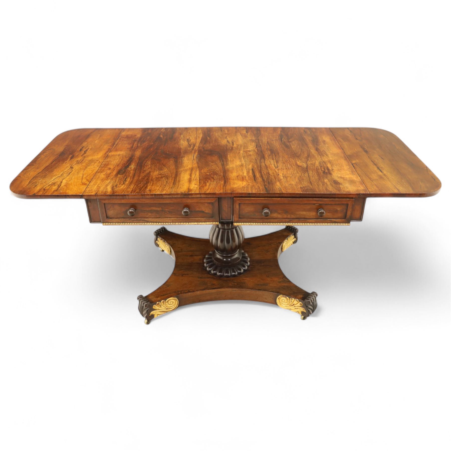 Regency rosewood sofa table, rectangular top with variegated rosewood veneers, hinged D-shaped leaves, over a set of dummy drawers to the back and full-size drawers to the front, with turned knob handles and gilt beaded moulding on frieze above elegant turned, tapering, and fluted, pedestal with a well-carved collar, supported on a large, tripartite concave platform base terminating in richly carved lion paw feet with gilded acanthus leaf carving, on concealed brass castors, circa 1815, length extended 161cm 
