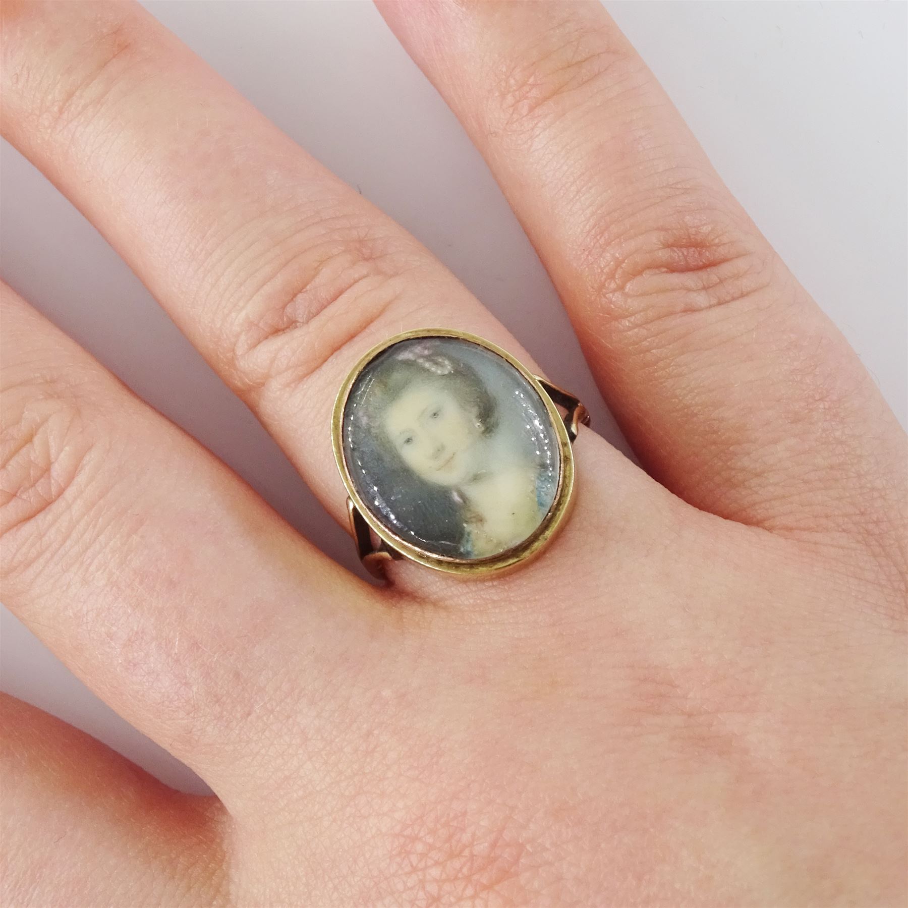 19th century gold portrait ring, set with an oval miniature of a woman wearing a pearl hairpin and earrings on ivory, under a glazed panel, set into a later gold ring with split shoulders. This lot has been registered for sale under Section 10 of the APHA Ivory Act
