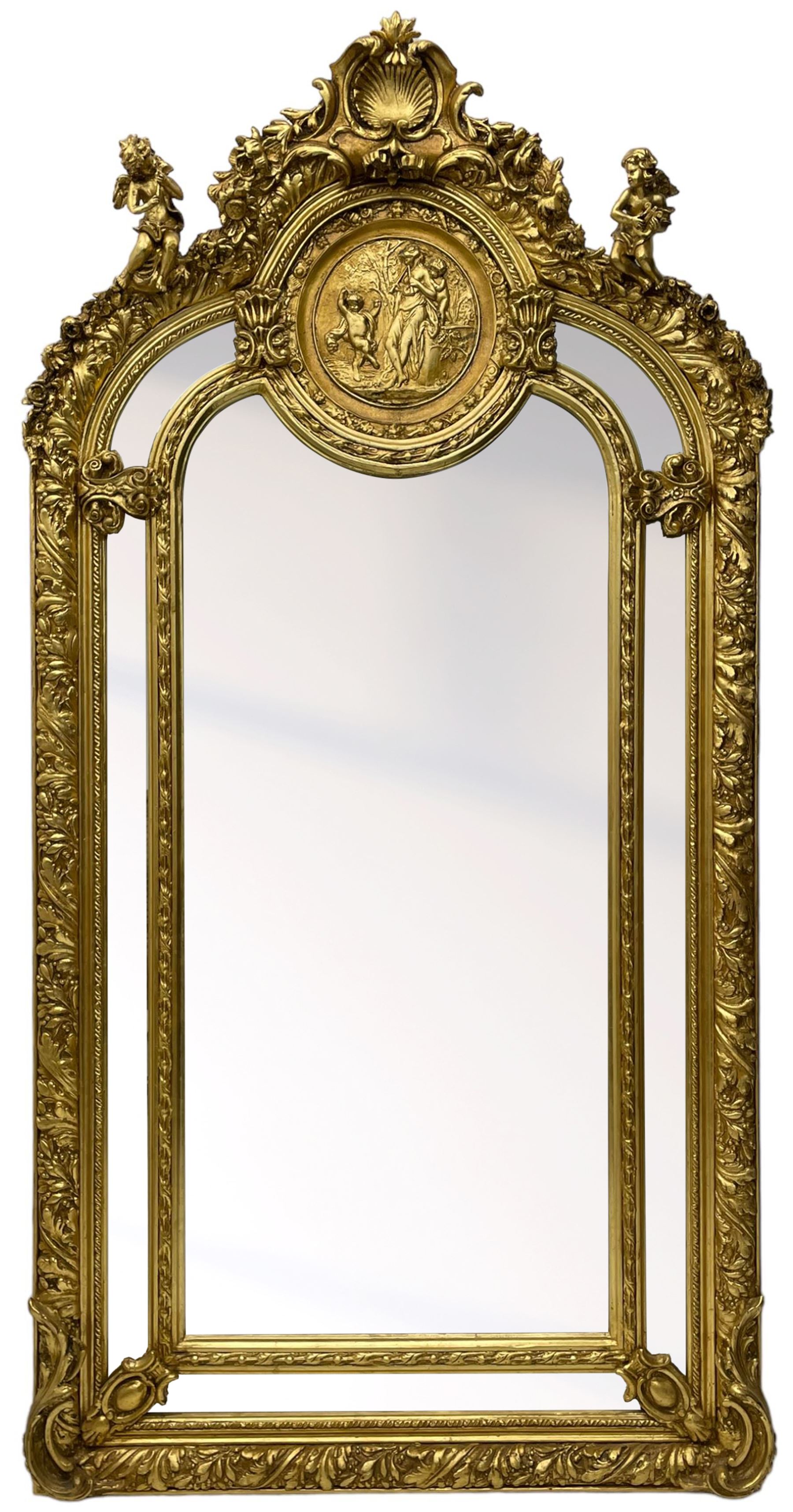 Large Italian Baroque design gilt wall mirror, the shell pediment over circular panel depicting classical female flute player with dancing putto within a tree landscape, the stepped arched upper frame surmounted by musical instrument playing putto and trailing flower heads, central bevelled mirror plate surrounded by plain segmented mirror plates, acanthus leaf moulded outer frame and foliate moulded inner slip