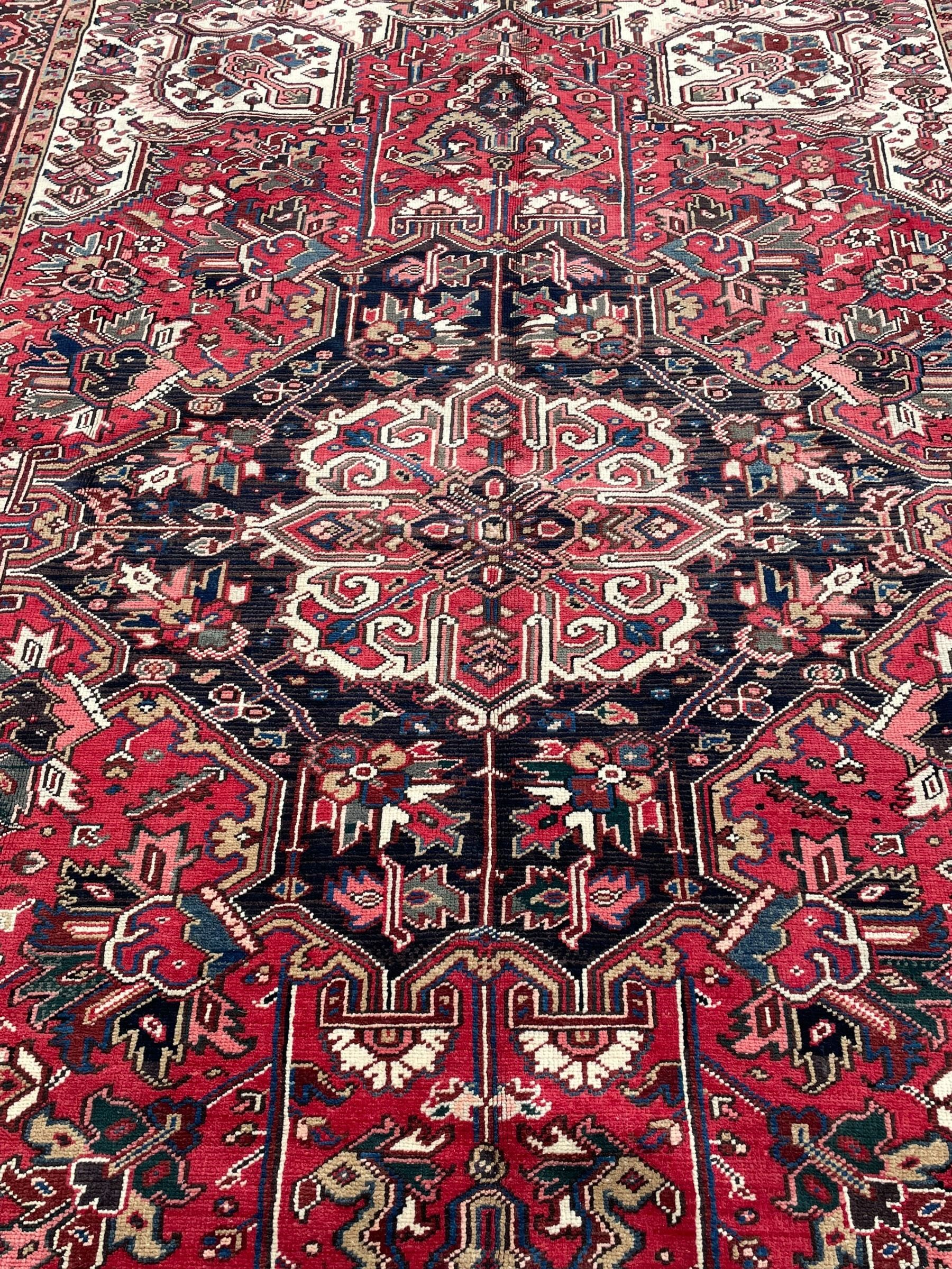 Persian Heriz red ground rug, central scrolled medallion on dark ground decorated with floral motifs, the surrounding field decorated with stylised plant motifs and smaller geometric symbols, guarded border with repeating pattern punctuated with rosettes and flower heads 