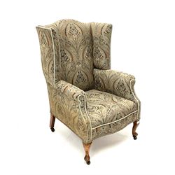 Early 20th century high wing back sprung armchair, upholstered in a stylised patterned fabric and trimming, cabriole supports and castors