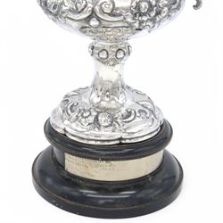 Victorian silver twin handled trophy, the bowl embossed with floral and scrolling decoration, with presentation engraving to body and twin acanthus capped C scroll handles, upon a lobed foot, hallmarked London 1866, maker's mark AM probably for Alexander Macrae, upon an ebonised wooden plinth, trophy only H25.5cm