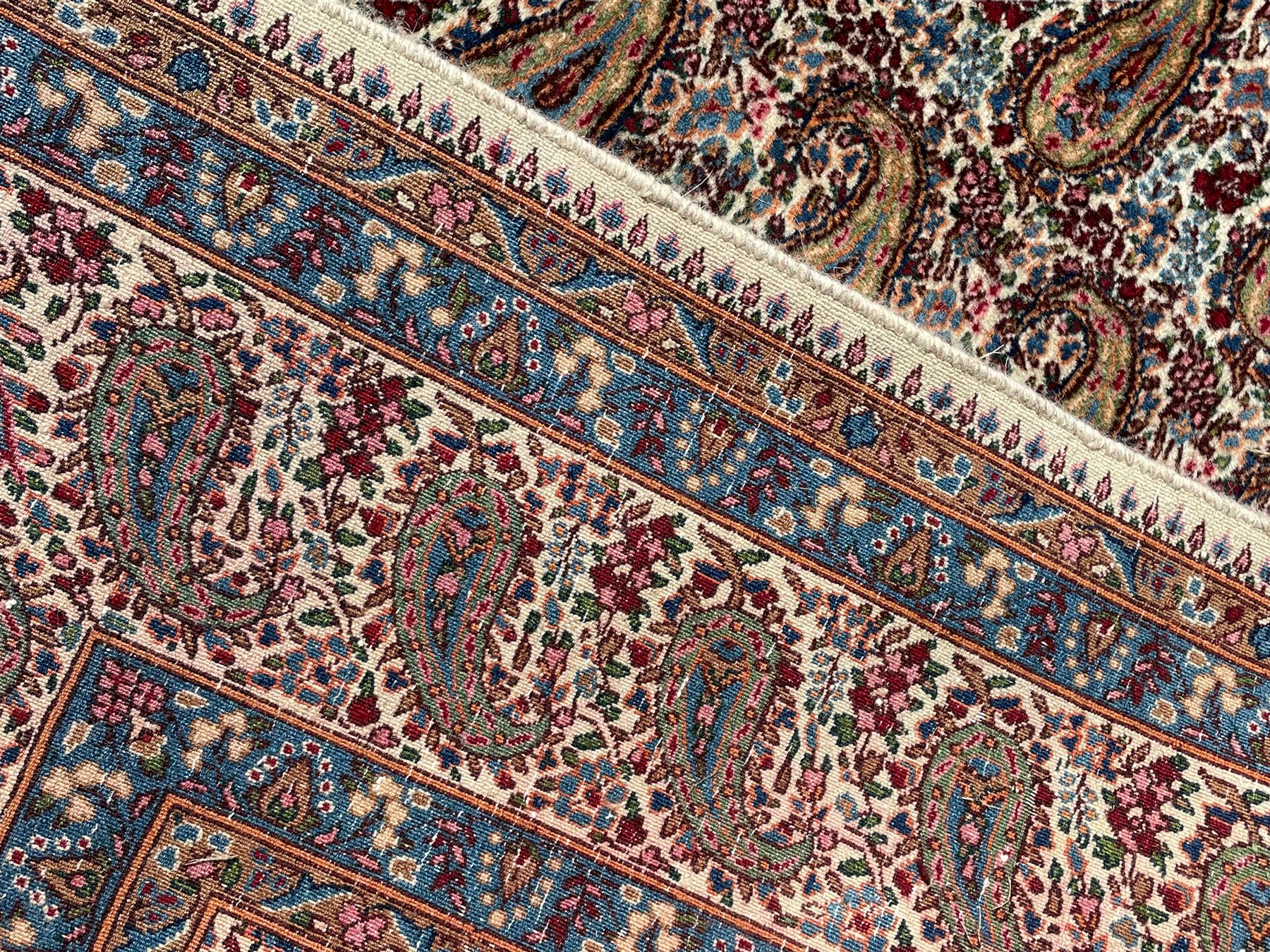 Persian Kirman carpet, the ivory ground field and border decorated with repeating Boteh motifs, interspaced with all-over floral design, blue ground border guards, the top border with signature panel