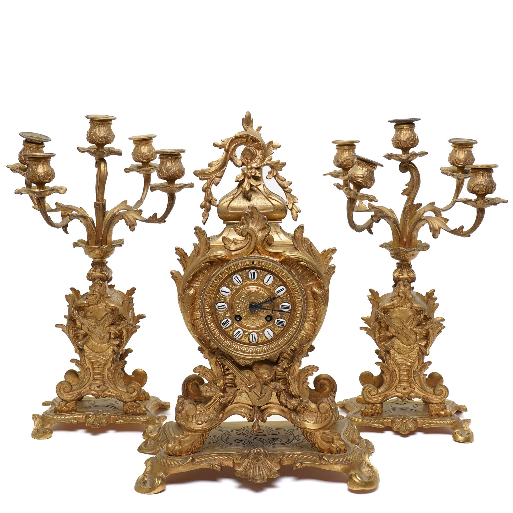 French-Late 19th century 8-day gilt brass rococo style mantel clock, c1890, waisted case  decorated with scroll work, leaves and musical instruments, resting on an attached plinth with splayed feet, dial within a glazed brass bezel with a gilt repoussé centre, porcelain cartouche Roman numerals and steel fleur-di-Lis hands, with a twin train movement, recoil anchor escapement and countwheel strike, sounding the hours and half-hours on a bell. With a matching pair of five-light candelabra. No Pendulum or Key.