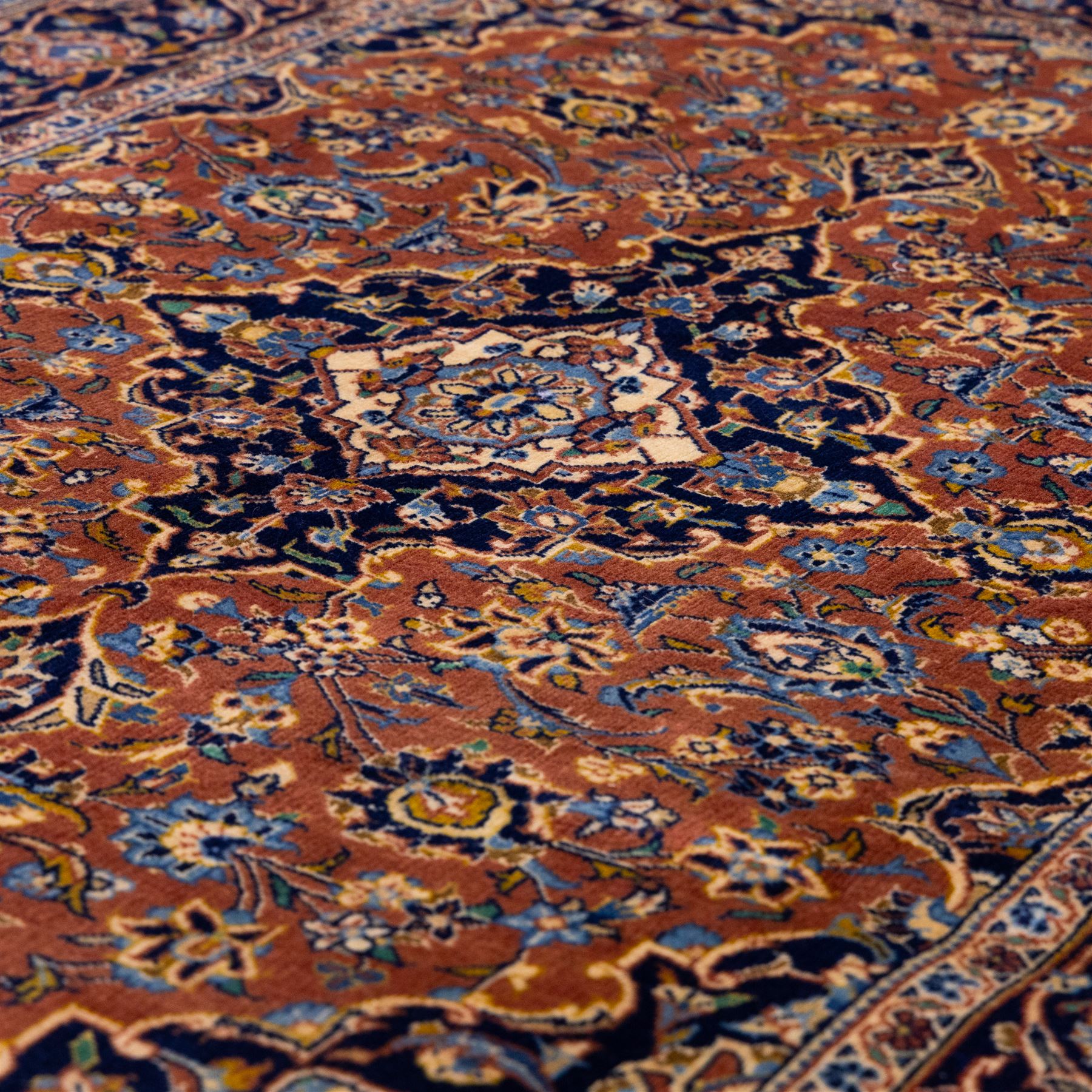 Central Persian Kashan hand-knotted rug,  terracotta field with central shaped ivory and dark blue medallion with palmettes, floral sprays and scrolling vine ornament, with conforming corner spandrels enclosed by a broad navy border of palmettes and flowering vine between multiple guard stripes, with cotton fringe ends 
