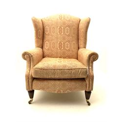 Late 20th century traditional shaped wingback armchair upholstered in pale pink patterned fabric, turned and fluted front supports with brass cups and castors