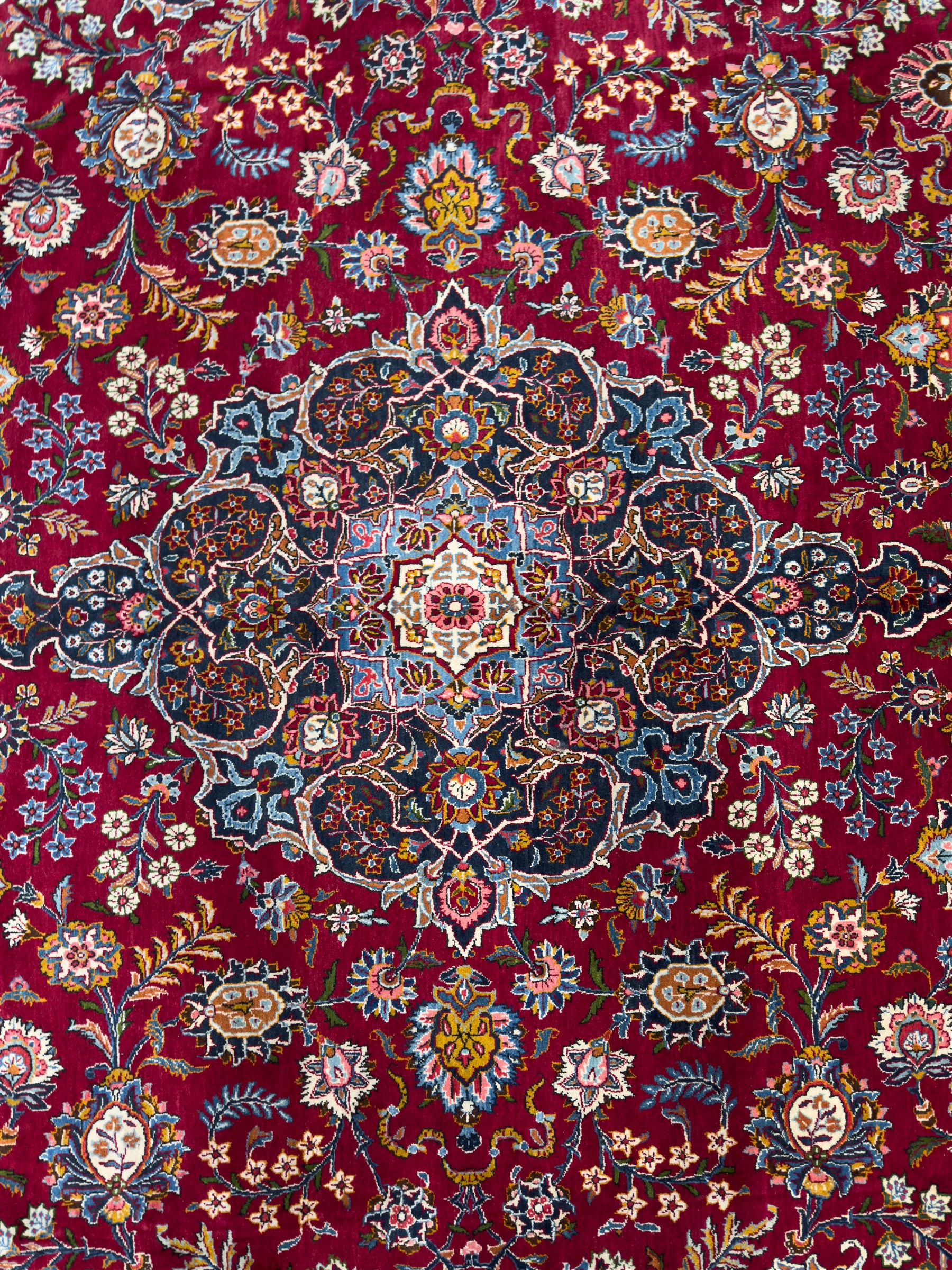 Central Persian Kashan hand-knotted carpet, madder red field with an intricate design of scrolling vines, palmettes, and rosettes with a central lobbed medallion with pendant motifs in shades of blue and ivory, and spandrels all within a blue-ground border of palmettes and flowerheads between multiple guard borders, 450cm x 335cm 