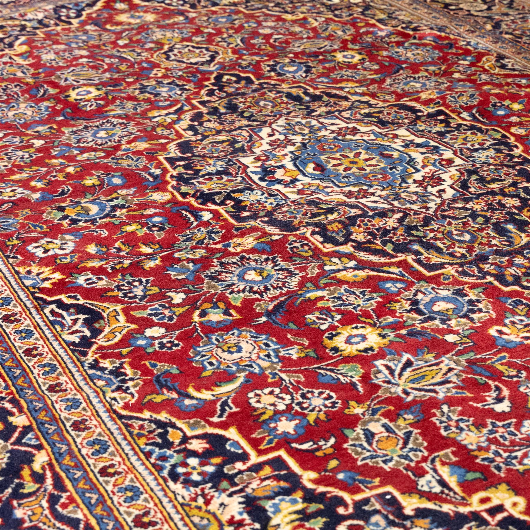 Central Persian Kashan hand-knotted rug, rich red field with a central lobbed medallion surrounded by finely drawn scrolling floral sprays, palmettes, and rosettes, enclosed by a broad dark blue floral border with multiple guard stripes and cotton fringe ends 