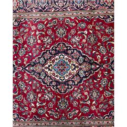 Central Persian Kashan hand-knotted carpet, the deep red ground with a central lozenge medallion in ivory and navy, surrounded by dense scrolling floral and vine motifs, navy spandrels and a wide border with stylised palmettes and flowerheads enclosed by multiple guard stripes