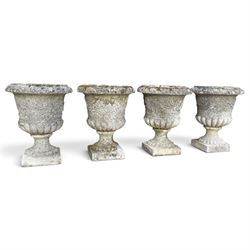 Set of four weathered composite stone urns of campana form, the everted leaf-moulded rolled rim over a body decorated with scrolling fruiting grapevines in relief, over a gadrooned underbelly and waisted socle, lobed collar on square base