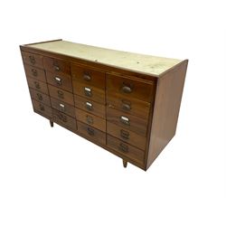 Mid-20th century teak haberdashery shop cabinet, sloped front and fitted with twenty drawers, metal cup handles and label holders, glass top with bass rule, on turned tapering feet