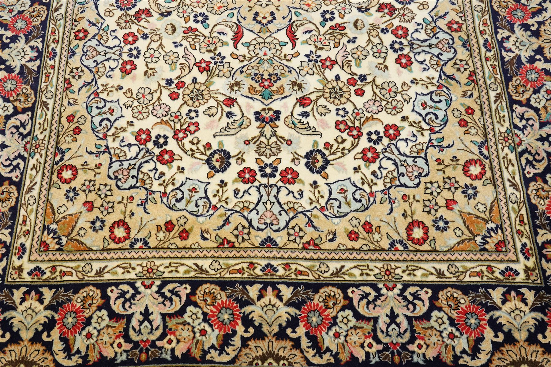 Central Persian Kashan hand-knotted carpet, ivory ground with a central floral medallion with an all-over design of scrolling vinery, palmettes, and flowering sprays i tones of madder red, cobalt, navy blue and sage green, with conforming spandrels, main border of deep indigo ground with repeating palmettes and stylized floral heads, flanked by narrow guard stripes, with cotton fringe ends, with cotton fringe ends 