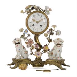 French- early 20th century 8-day Chinoiserie brass and porcelain mantel clock in the 18th century style, brass base with splayed feet profusely decorated with clusters of painted porcelain flowers and two decoratively painted Chinese foo dogs, movement enclosed in a circular porcelain movement case illustrated with birds of paradise and oriental motifs, with a white enamel dial Arabic numerals, minute markers, pierced gilt hands and a floral swag, rack striking Parisian movement striking the hours and half-hours on a bell. With a visible sunburst pendulum and key.