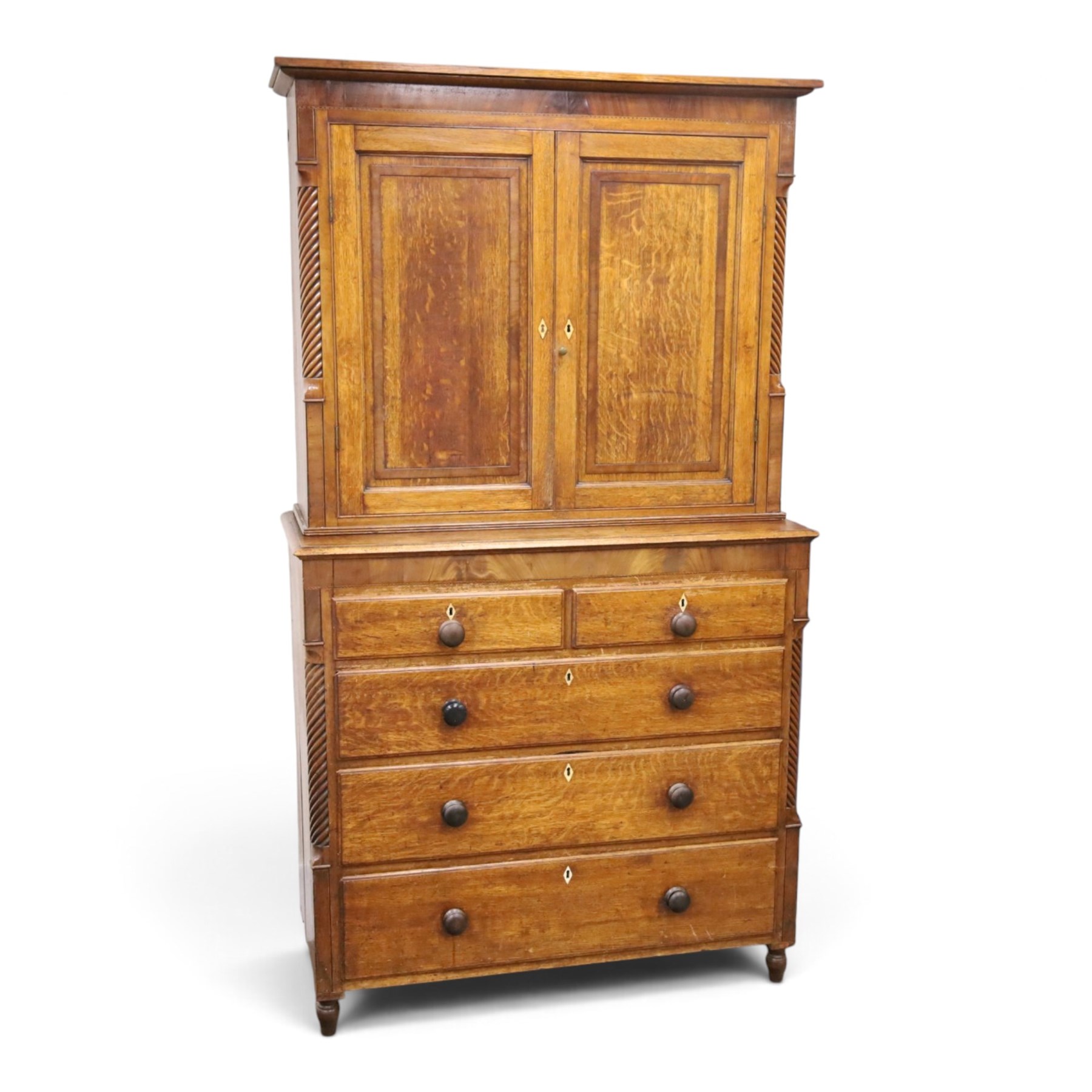 Georgian oak and mahogany housekeepers cupboard, the upper section with projecting cornice, crossbanded and inlaid frieze, above panelled doors with bone escutcheons, enclosing three fixed shelves, flanked by barley twist quarter pilasters, the lower section with further crossbanded and inlaid frieze, two short over two long graduating drawers with ebonised handles, raised on turned supports 