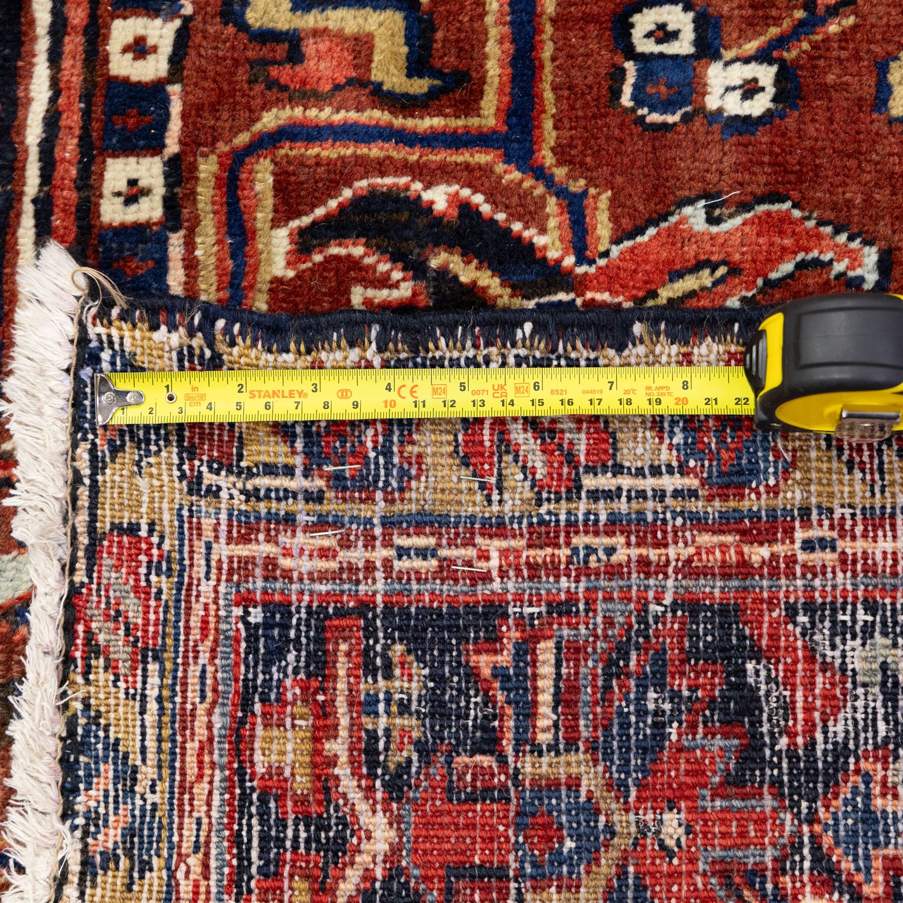 North West Persian Heriz hand-knotted carpet, madder red field, with a large angular stepped central medallion with serrated outlines, flanked by bold stylized palmettes, hooked leaves and geometric devices, spandrels similarly decorated within a wide geometric main border featuring angular floral motifs, enclosed by multiple guard borders in madder red, indigo blue, ivory, olive green and soft ochre, with cotton fringe ends 