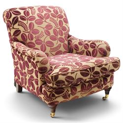 Traditional Howard shape armchair, rolled back and arms over deep seat, raised on turned supports with brass cups and castors, upholstered in plum foliate patterned fabric with loose seat cushion