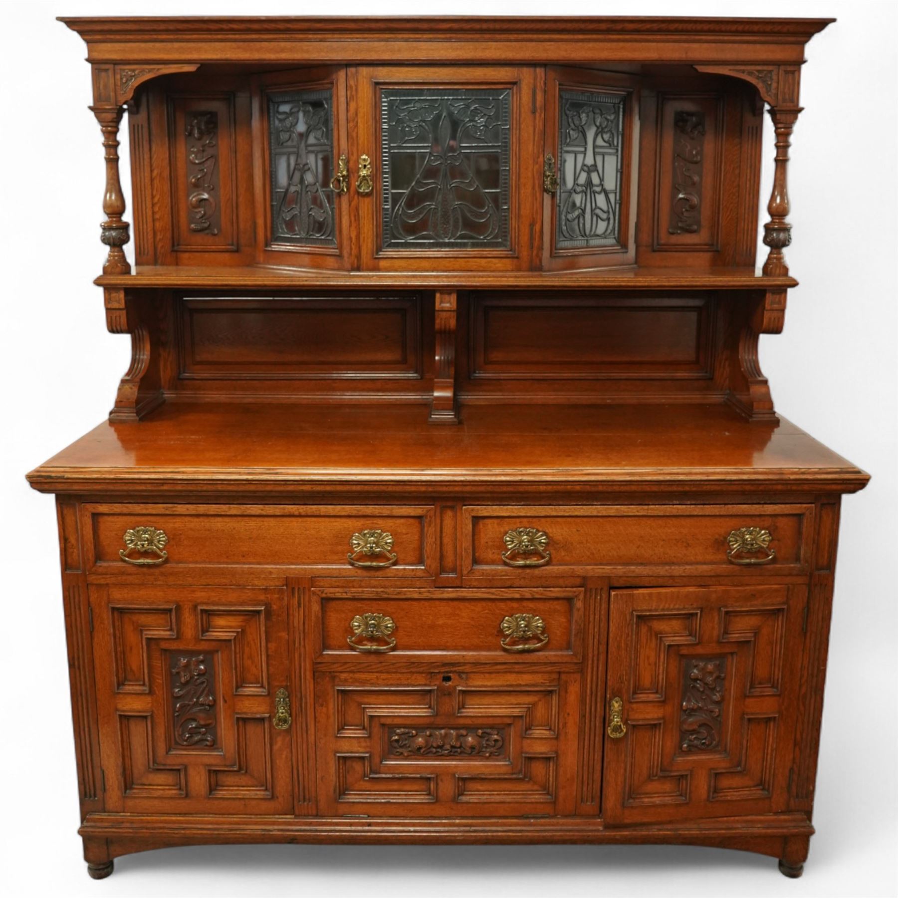 Early 20th century golden oak dresser sideboard, superstructure with three stained and leaded glass doors flanked by carved uprights above an open display shelf, the base with three drawers and geometric panelled cupboard doors, enclosing drawers with cutlery and bottle compartments, raised on short feet