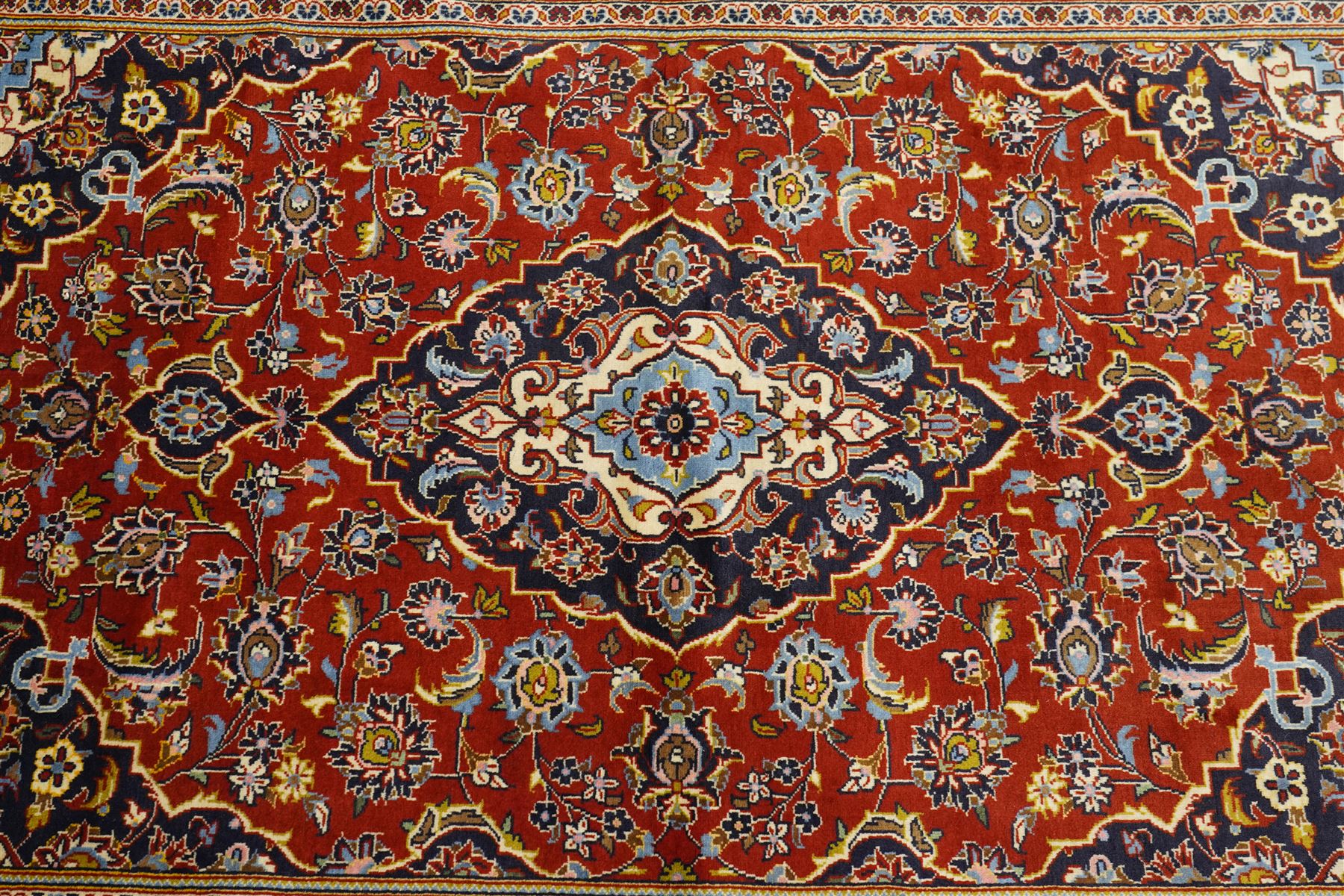 Central Persian Kashan hand-knotted rug, madder red field with central articulated cusped medallion in ivory and sky blue, issuing network of scrolling arabesques palmettes and floral sprays with conforming spandrels, indigo main border of scrolling vinery and stylized palmettes, framed by multiple guard borders, with cotton fringe ends  