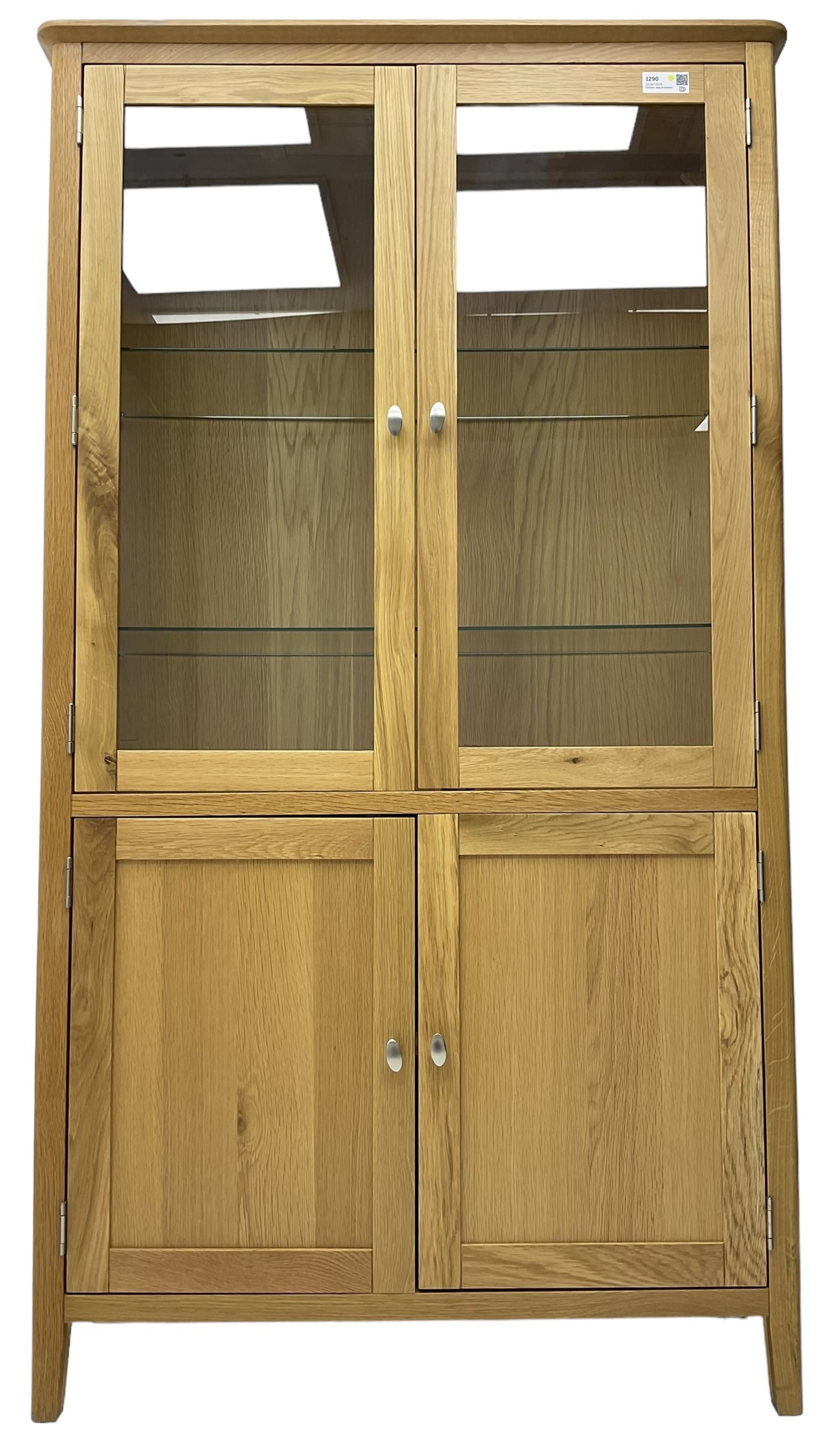 Contemporary light oak display cabinet on cupboard, fitted with two glazed doors enclosing two adjustable glass shelves with light fittings to the top, over panelled cupboard concealing single shelf, on square supports