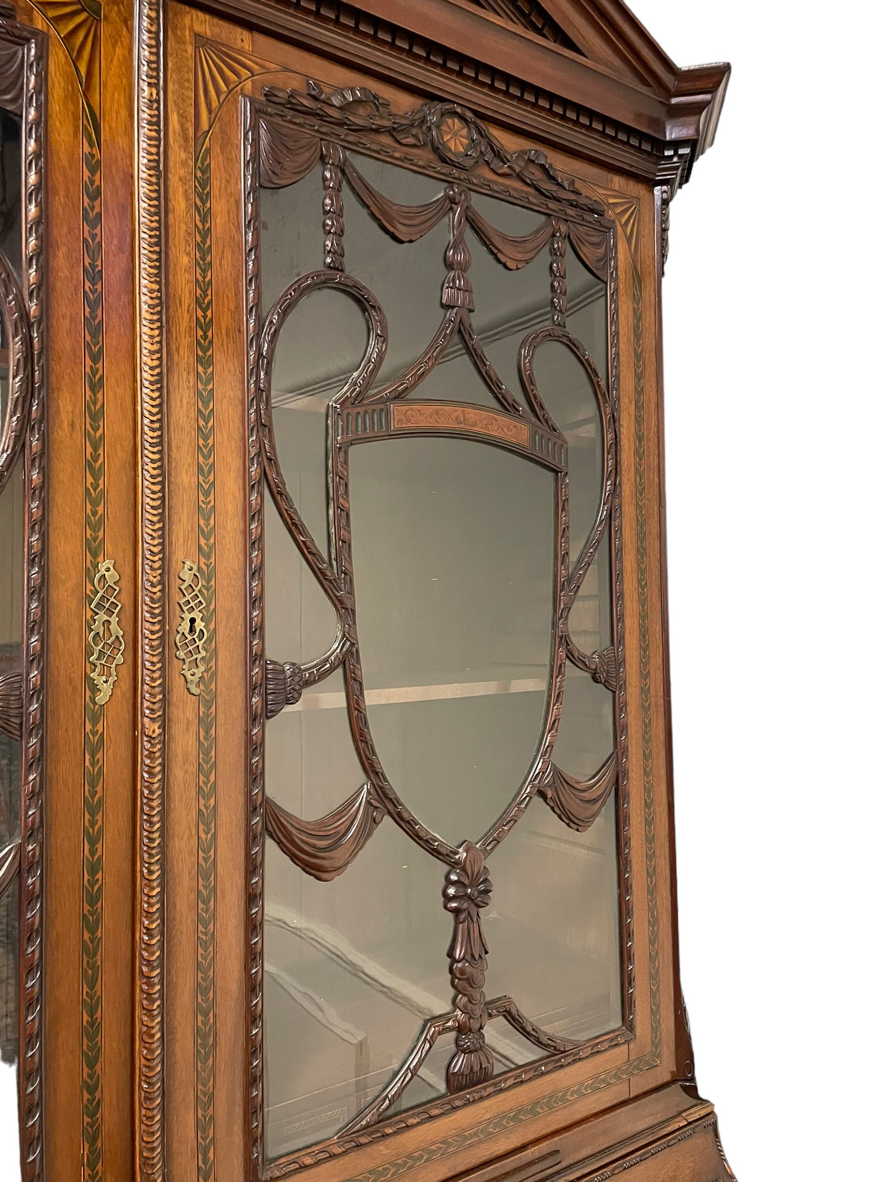 Large and impressive late 19th century Irish mahogany secretaire bookcase, moulded sloped arch dentil pediment decorated with central carved mask surrounded by linen festoons and trailing bellflowers, canted and fluted uprights with carved acanthus leaf upper and lower brackets, enclosed by two astragal glazed doors in the form of trophies with twist ribbon carved mouldings, inlaid with fanned spandrels and trailing leaf painted satinwood band, cylinder roll top inlaid with central fan motif and fanned spandrels, pull-out action revealing stepped fitted interior, central tambour roll cupboard flanked by two Composite pilasters and a combination of drawers and pigeon holes, fitted with three break-front drawers, matched canted and fluted corners with acanthus leaf carvings, on square tapering and fluted spade feet