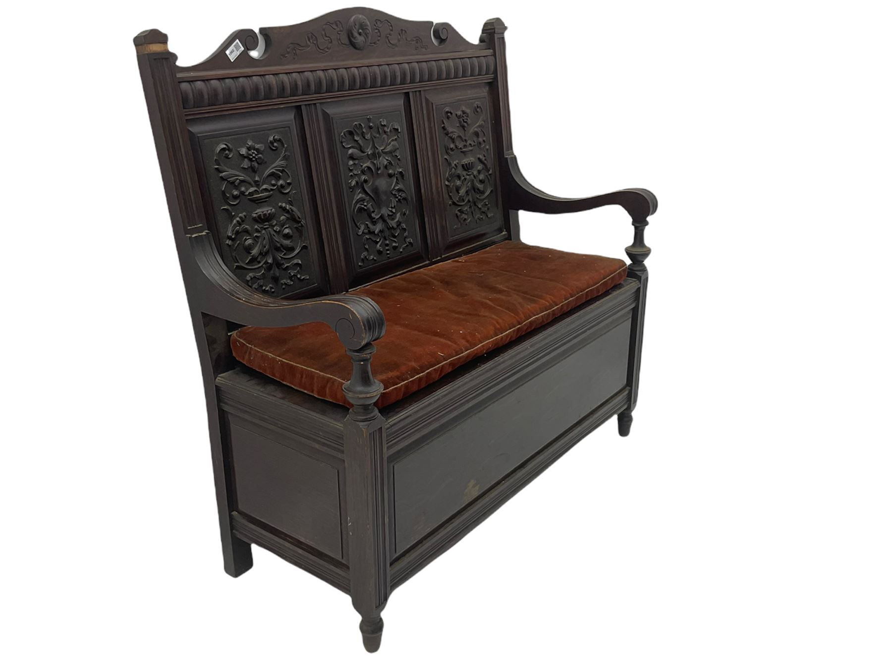 Late 19th century oak settle bench, shaped cresting rail carved with ribbon over triple panelled back, the panels relief carved with scrolled and floral urns and cartouche, hinged box seat, turned front feet, with upholstered squab cushion