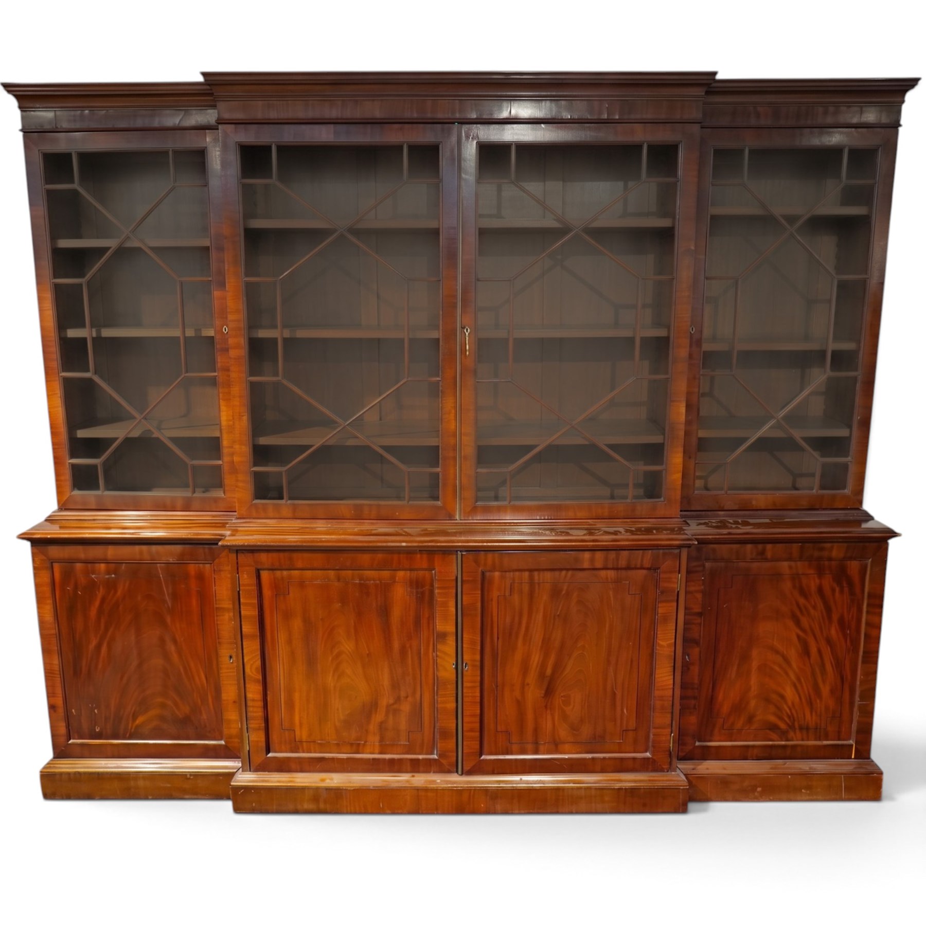 Fine Regency mahogany breakfront library bookcase, the stepped and moulded cornice over four astragal glazed doors enclosing adjustable shelves, the projecting central lower section with a pair of panelled doors enclosing three graduated drawers with brass inset handles above two deep zinc-lined cellarette compartments, flanked by panelled cupboard doors enclosing shelves, the figured mahogany fronts with ebony line inlay, on moulded plinth bases