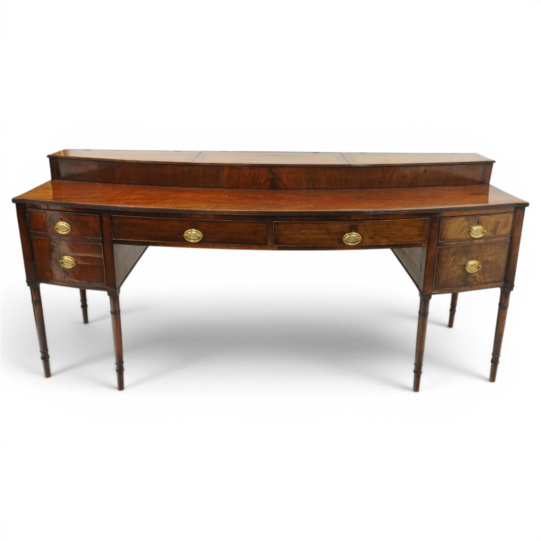 Large George III mahogany bowfront sideboard, the shaped top with raised superstructure fitted with three compartments with hinged tops and reeded edge, the body fitted with two central cock-beaded frieze drawers, flanked by a cellarette drawer and hinged cupboard both with faux double drawer facias, raised on ring turned tapering supports
Provenance: Kirkham Hall